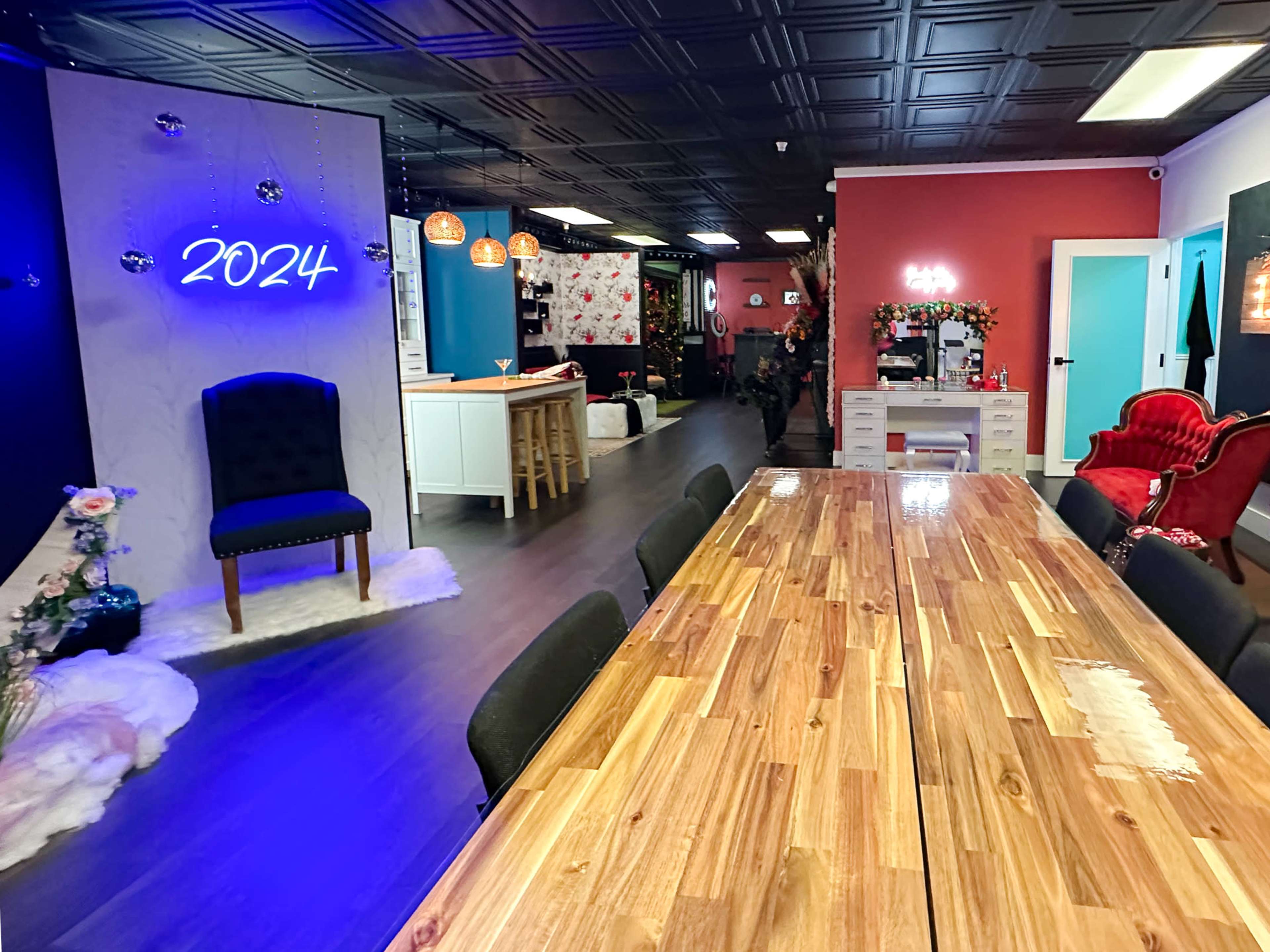 The image shows a spacious, modern interior with a large wooden table, a blue-lit wall displaying "2024," and various seating arrangements in a brightly colored setting.