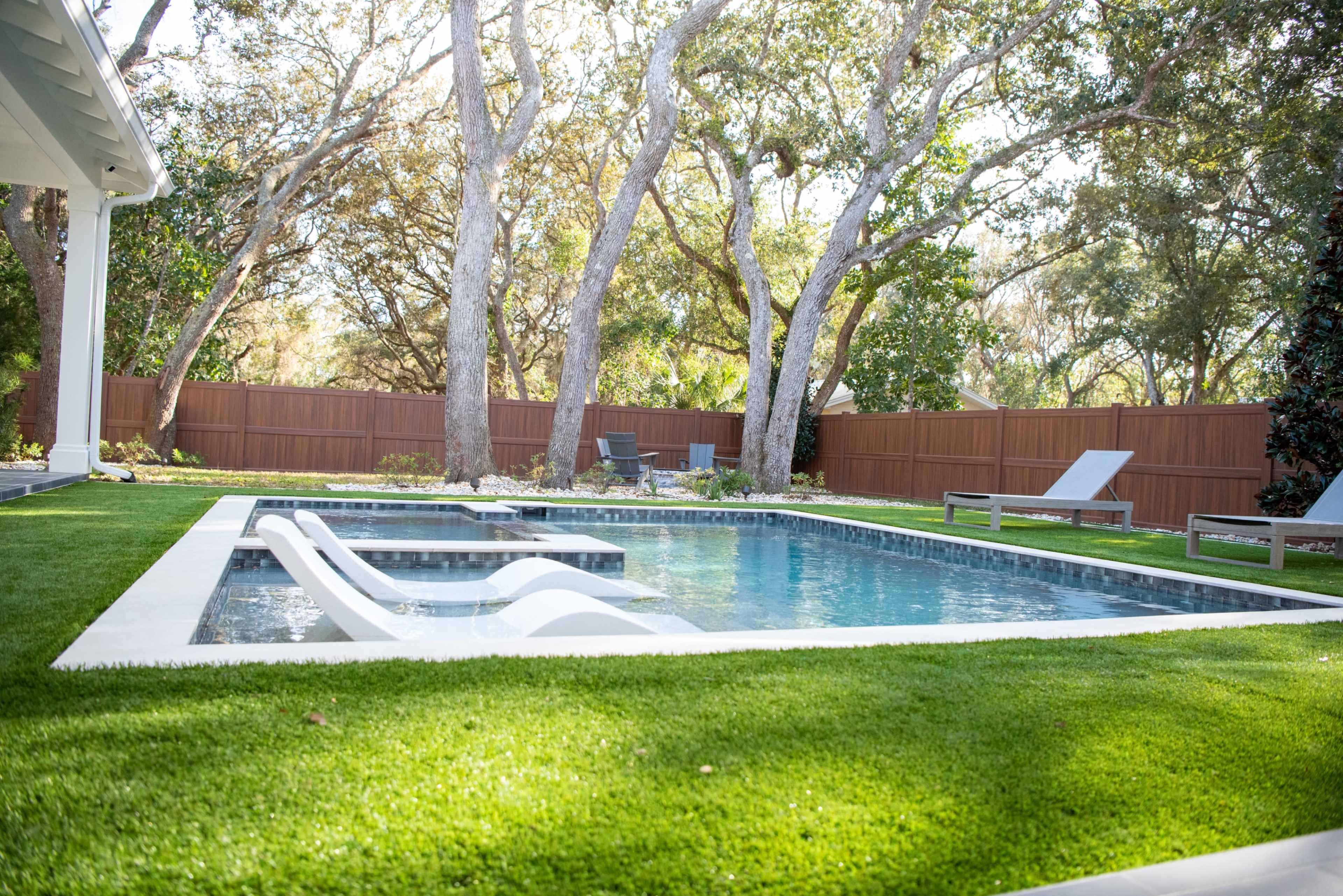 The image shows a rectangular swimming pool surrounded by artificial grass and trees in a backyard setting.