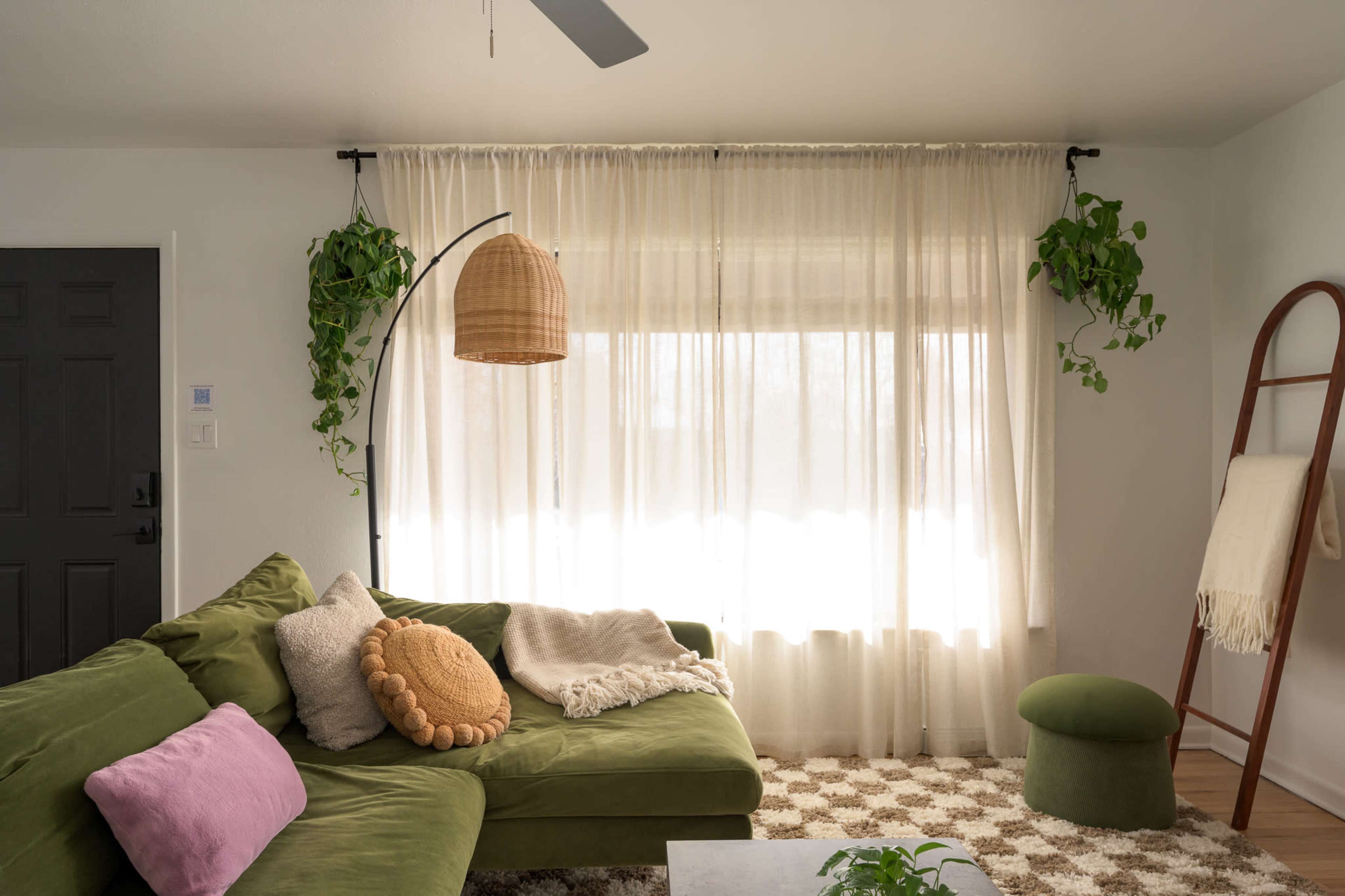 The living room features a green sectional sofa, a large window covered with sheer curtains, potted plants, and a ladder displaying a blanket.
