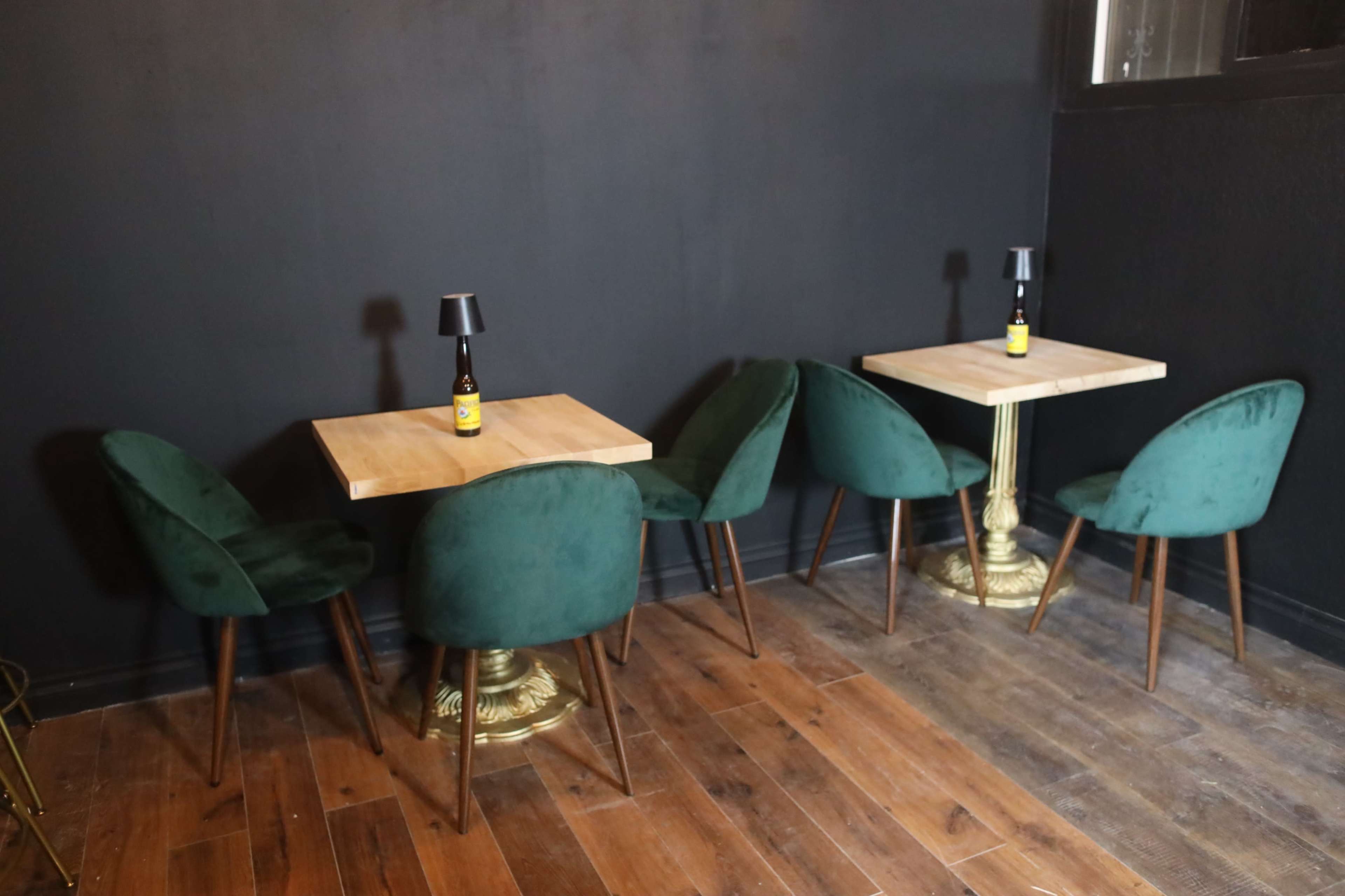 Two small tables with green velvet chairs are arranged on a wooden floor against a black wall, each topped with a small lamp and a bottle.