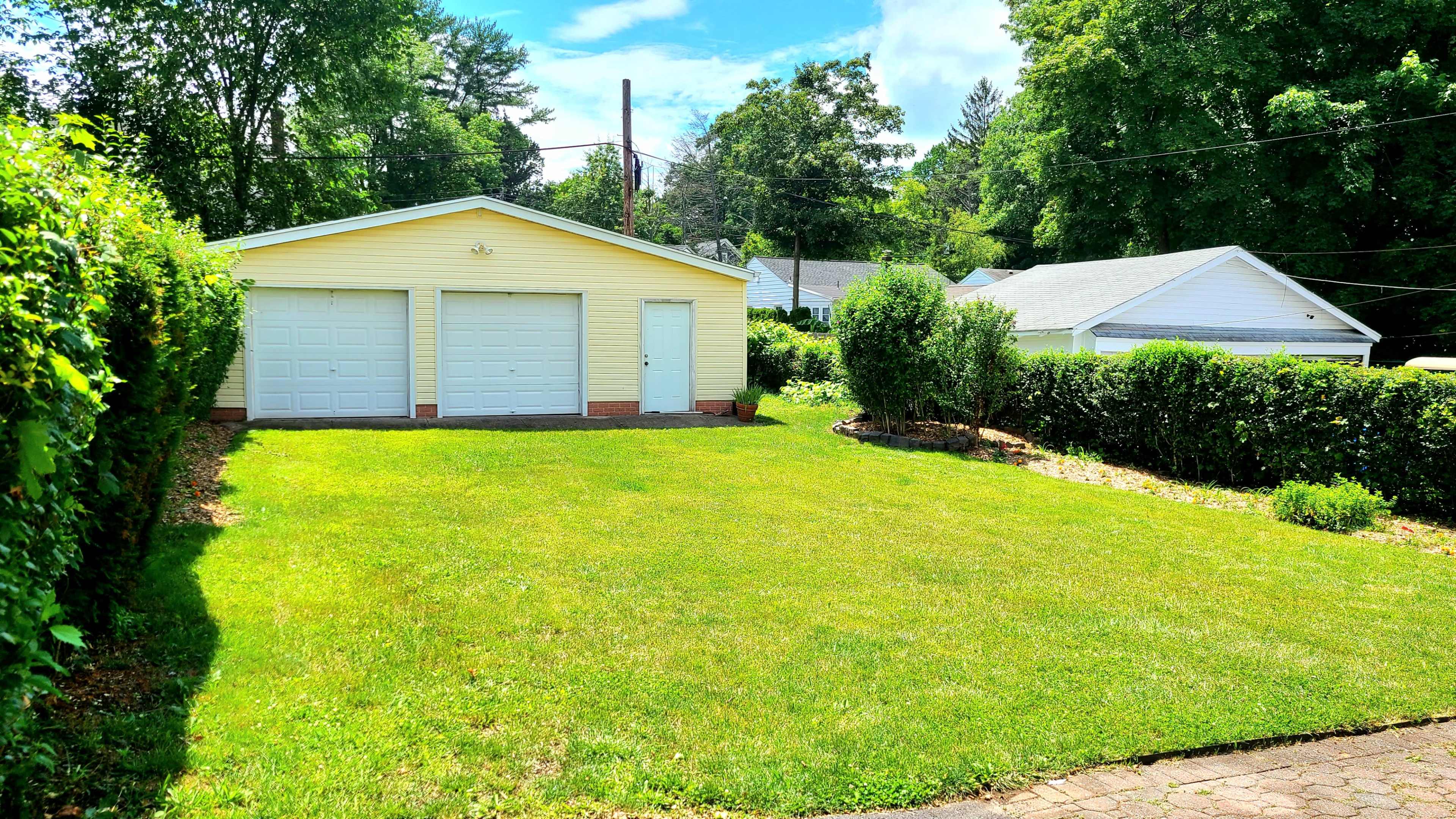 A two-car garage with a light yellow exterior is set in a grassy area surrounded by hedges, with additional buildings visible in the background.