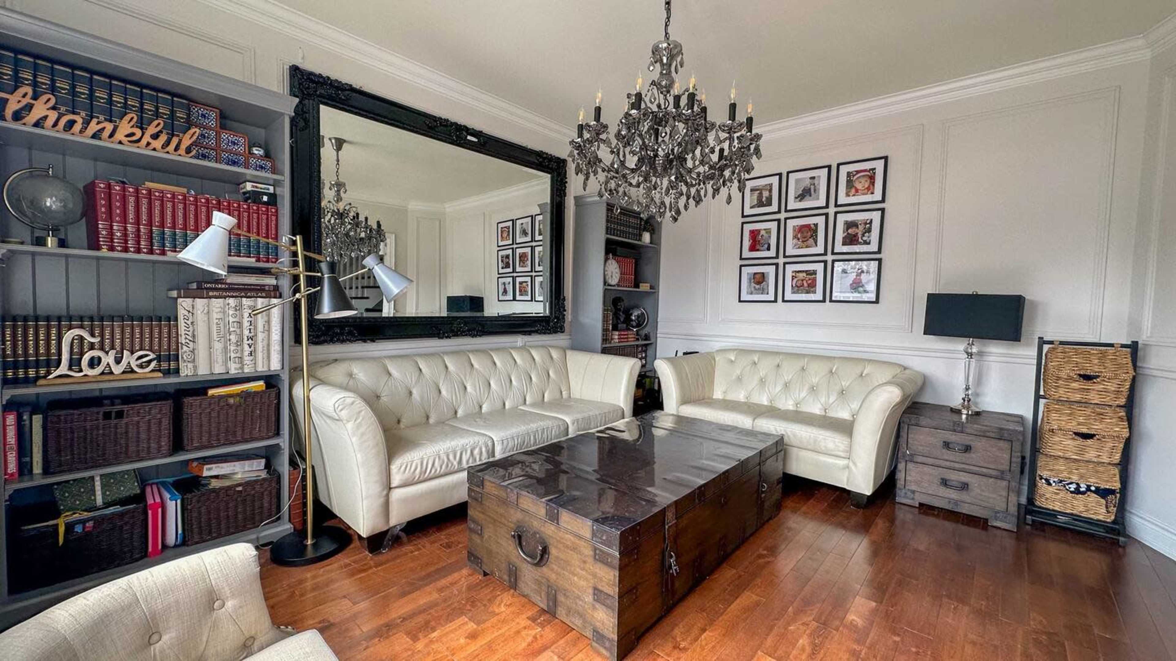 A well-furnished living room with two white leather sofas facing a wooden coffee table, surrounded by bookshelves and framed photographs.