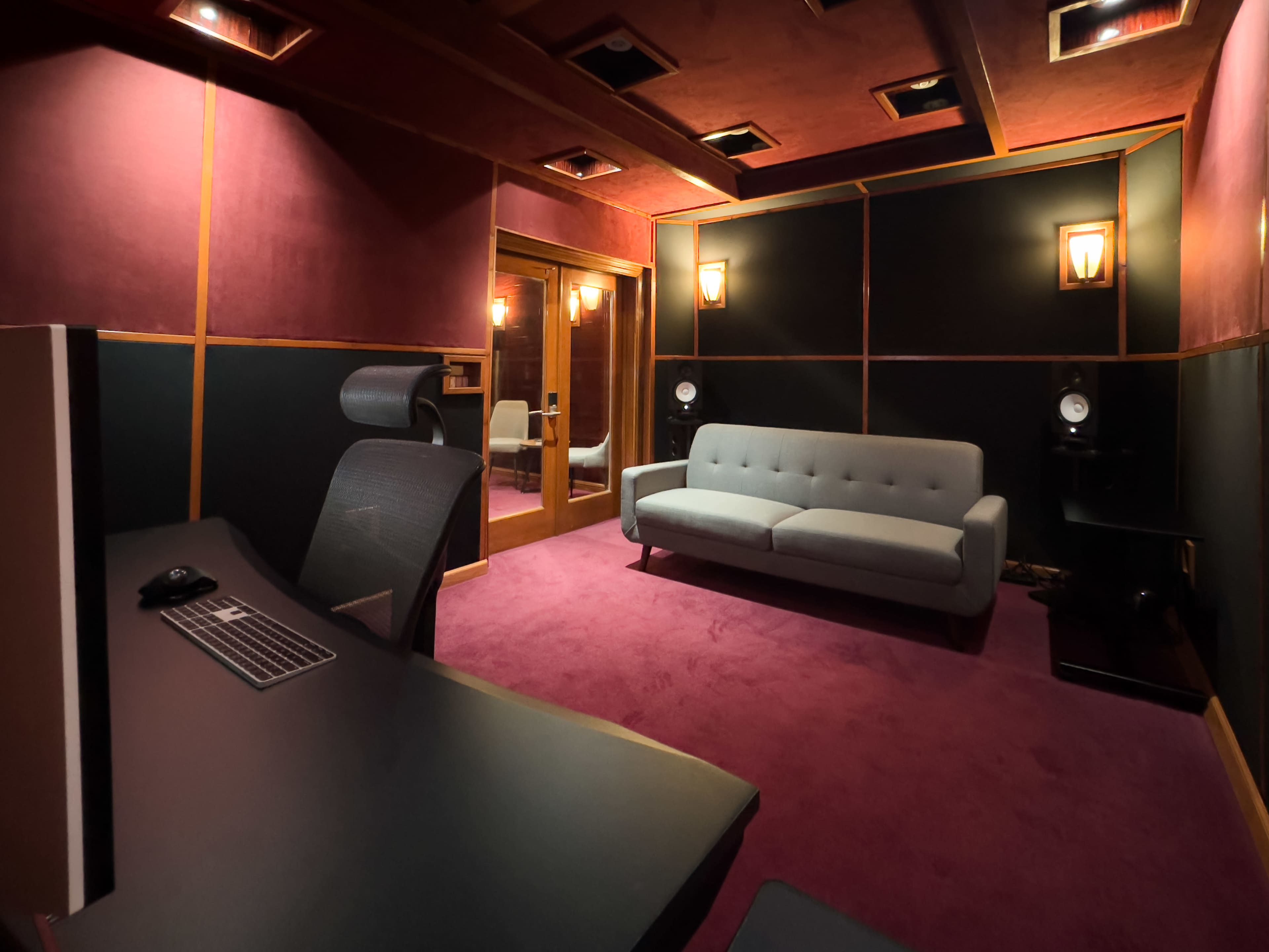 A sound studio with a desk, a gray sofa, and sound equipment, all situated in a room with dark walls and purple carpeting.