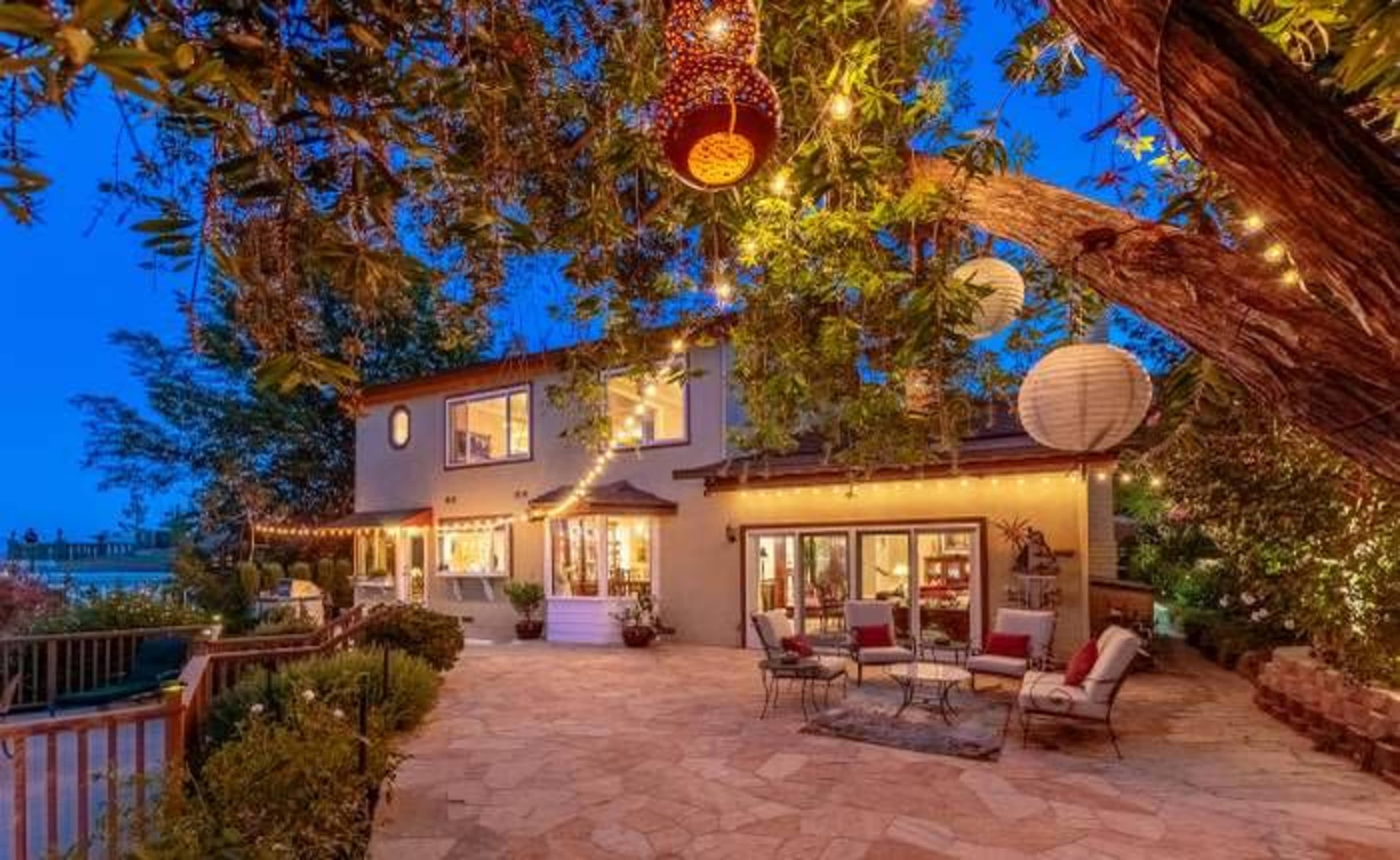 A two-story house with outdoor seating is illuminated by decorative lights and lanterns under a tree during twilight.