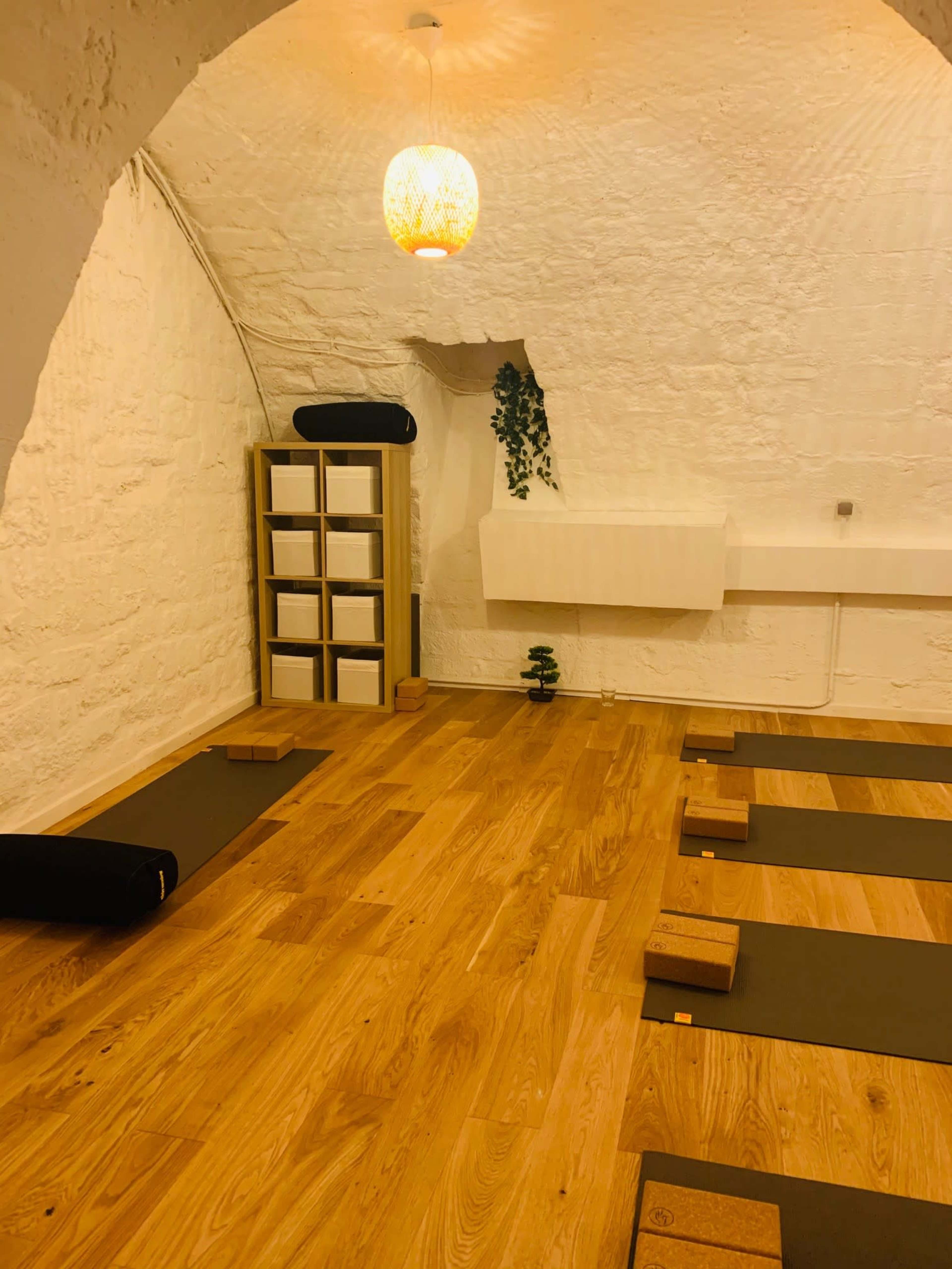 A yoga studio with wooden floors, neatly arranged mats, and a shelf filled with white storage boxes under an arched ceiling.