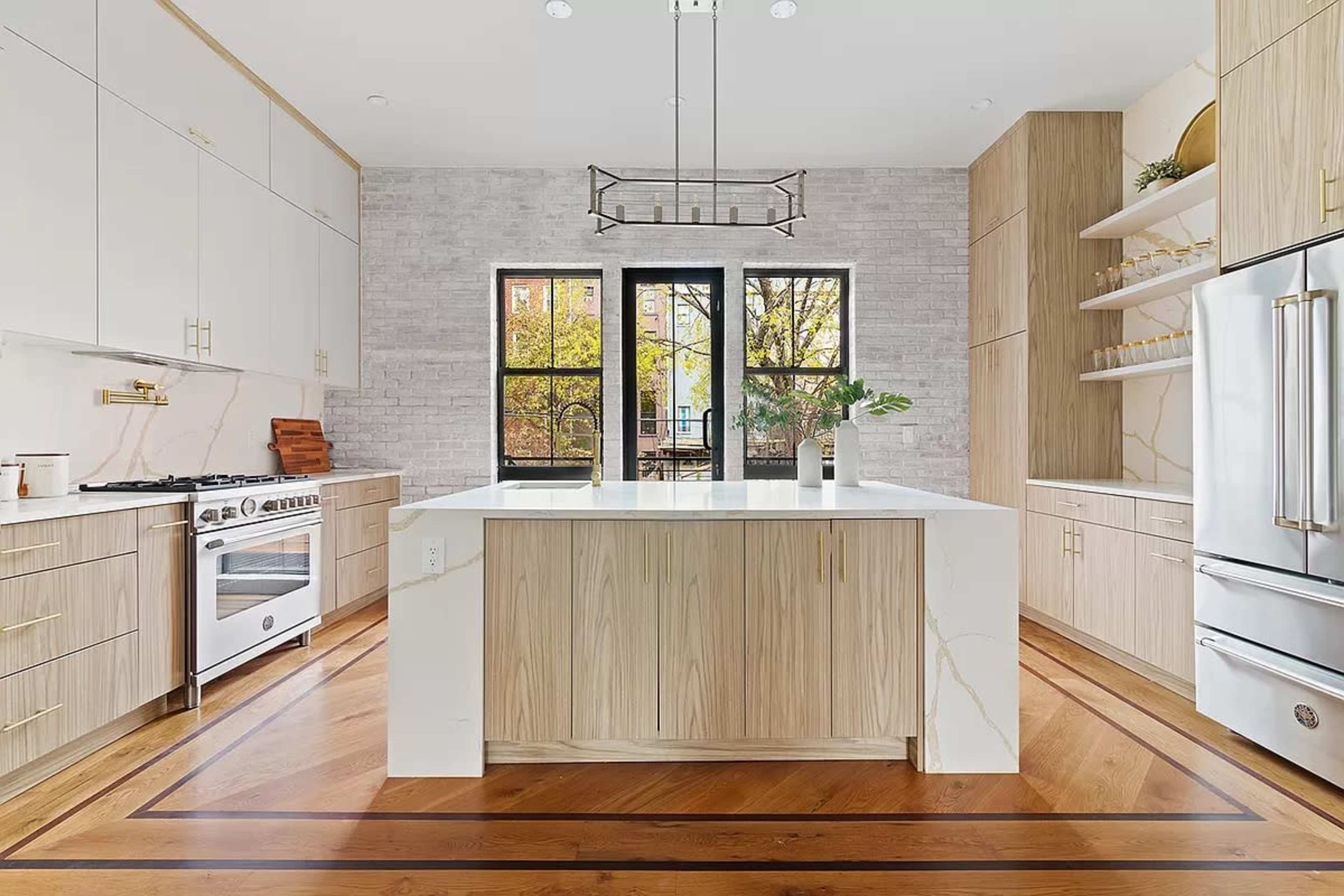 The kitchen features a large central island with a white countertop, wooden cabinetry, and modern appliances against a backdrop of brick walls and large windows.