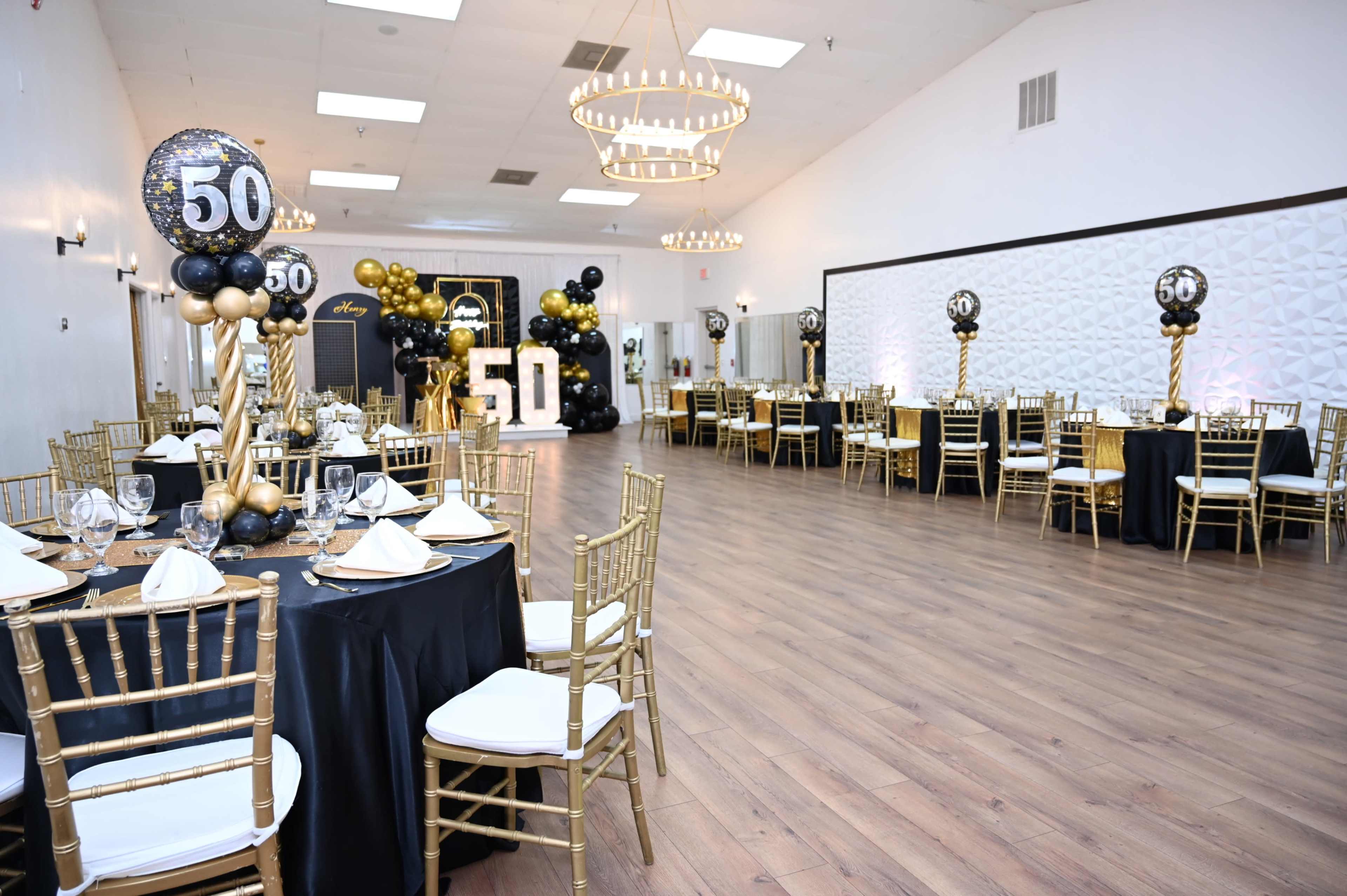 The image shows a spacious event hall decorated for a 50th birthday celebration, featuring black and gold table settings, elegant chairs, and decorative balloons.