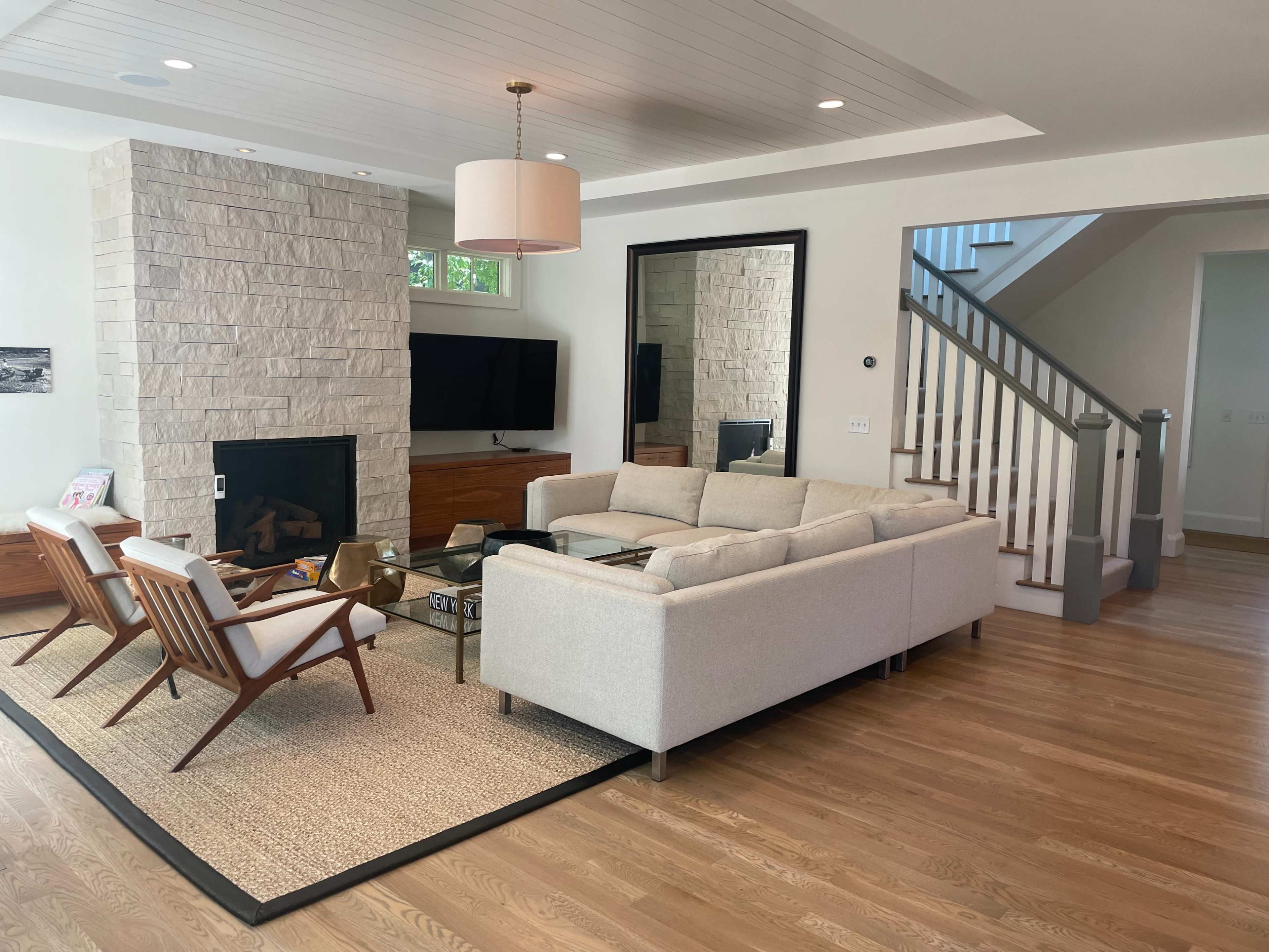 The image shows a modern living room with a light-colored sofa, a stone fireplace, a television mounted on the wall, and a staircase in the background.