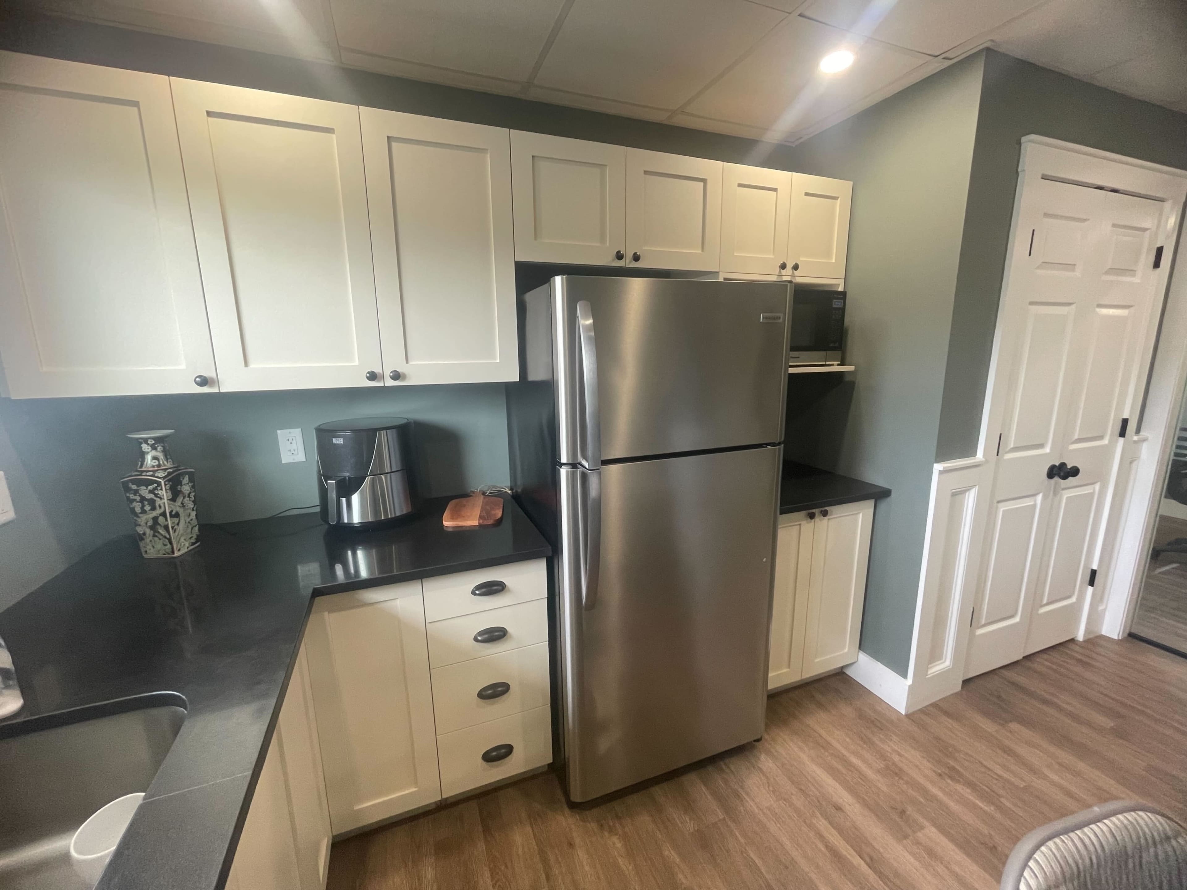 The image shows a modern kitchen with light-colored cabinets, a stainless steel refrigerator, and a countertop along with a coffee maker and microwave.