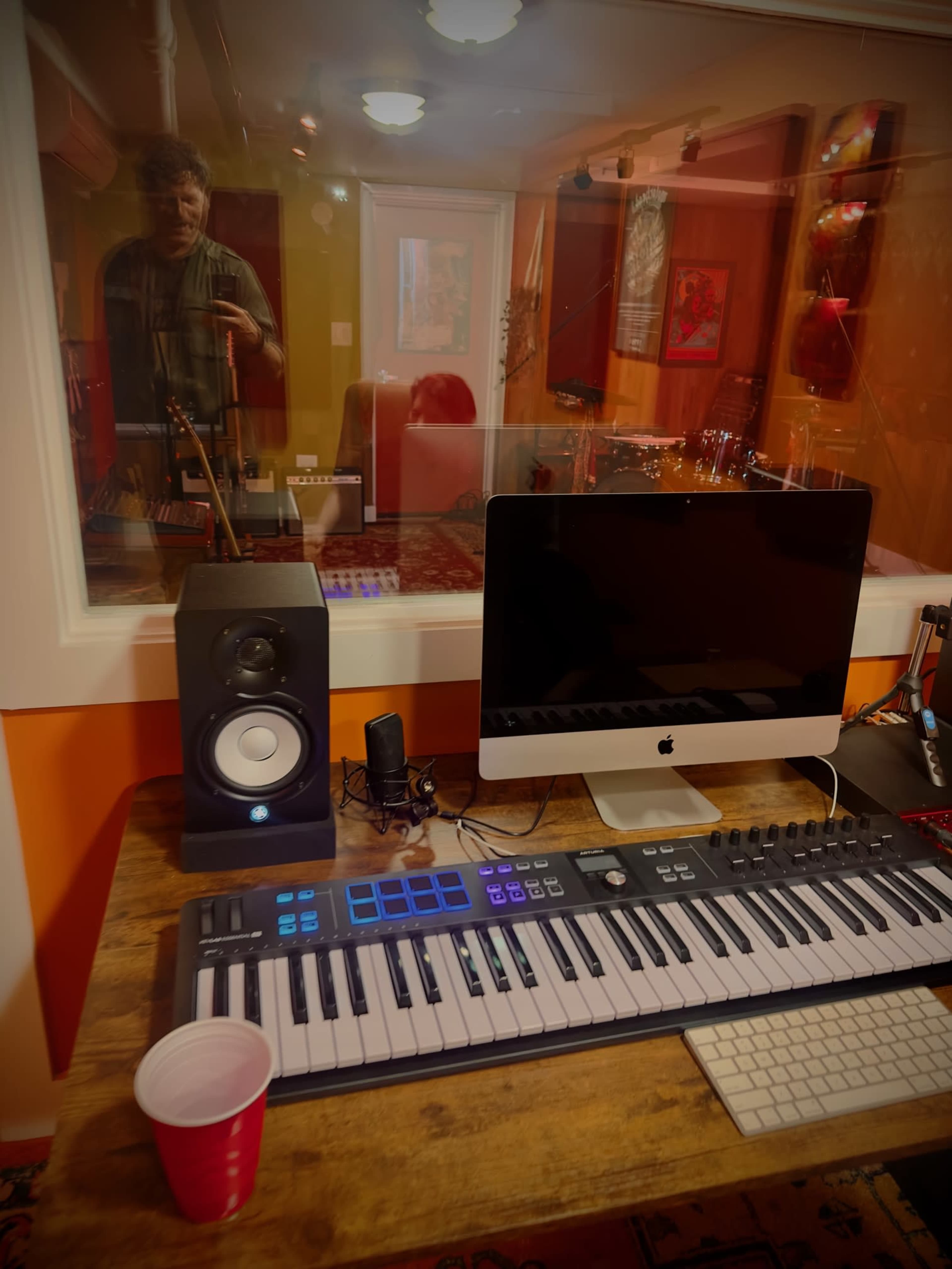 A music production studio with a keyboard, monitor, and audio equipment on a desk, and a person reflected in the glass window.