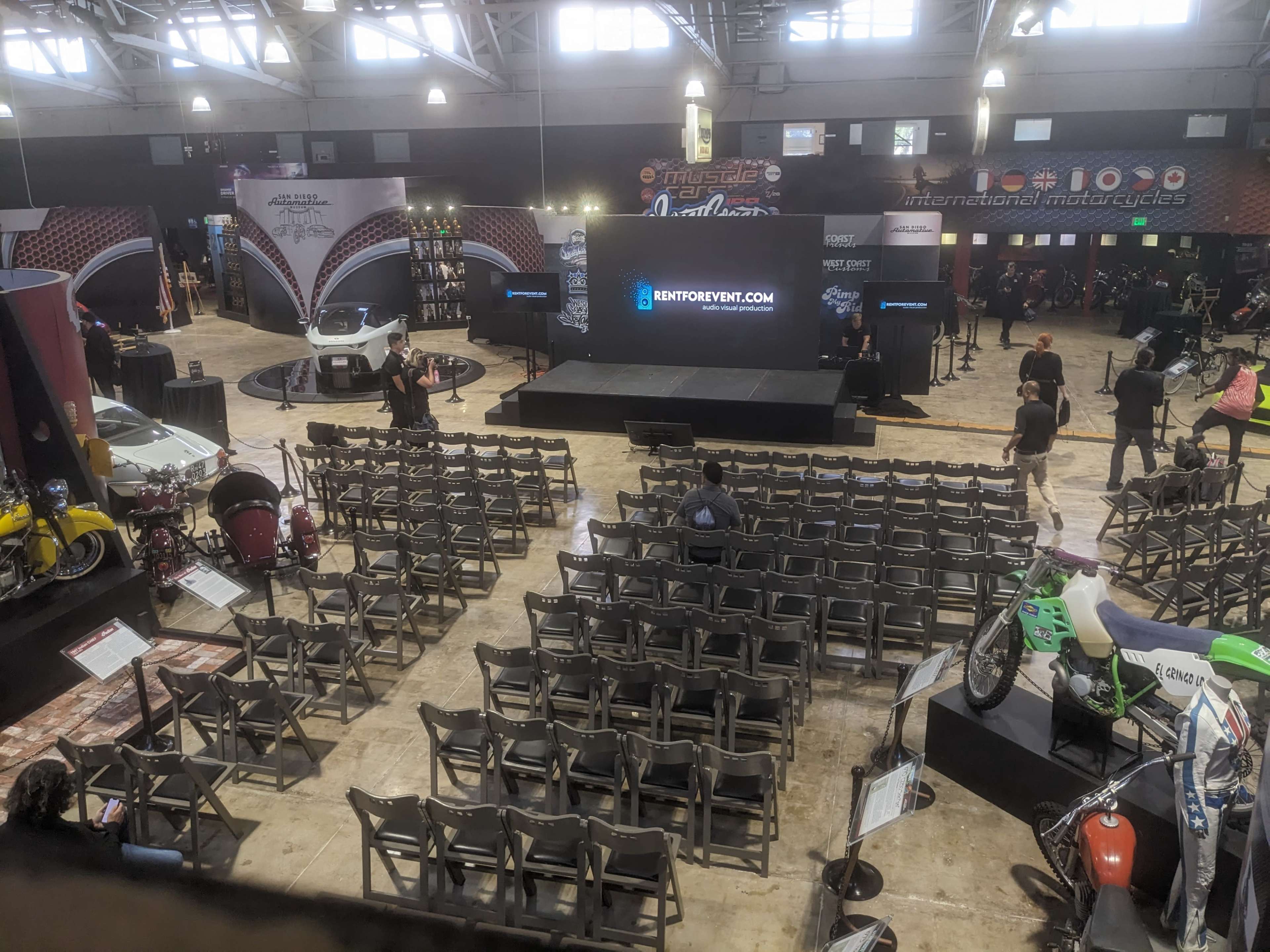 The image shows an event venue set up for a presentation, featuring empty rows of chairs facing a stage with a large screen, surrounded by various motorcycle displays.