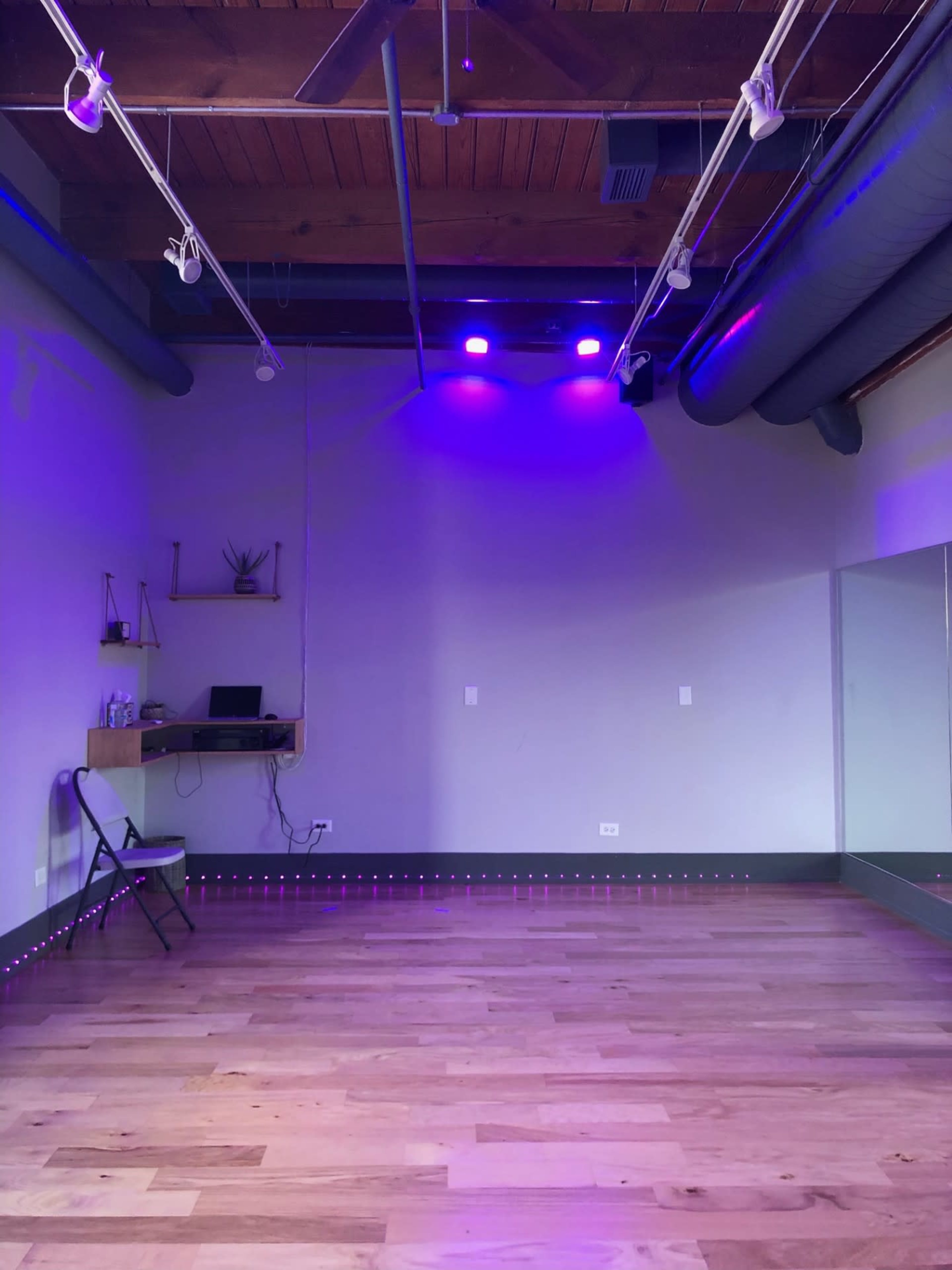 An empty dance studio features wooden floors, a wall-mounted mirror, and purple LED lights.