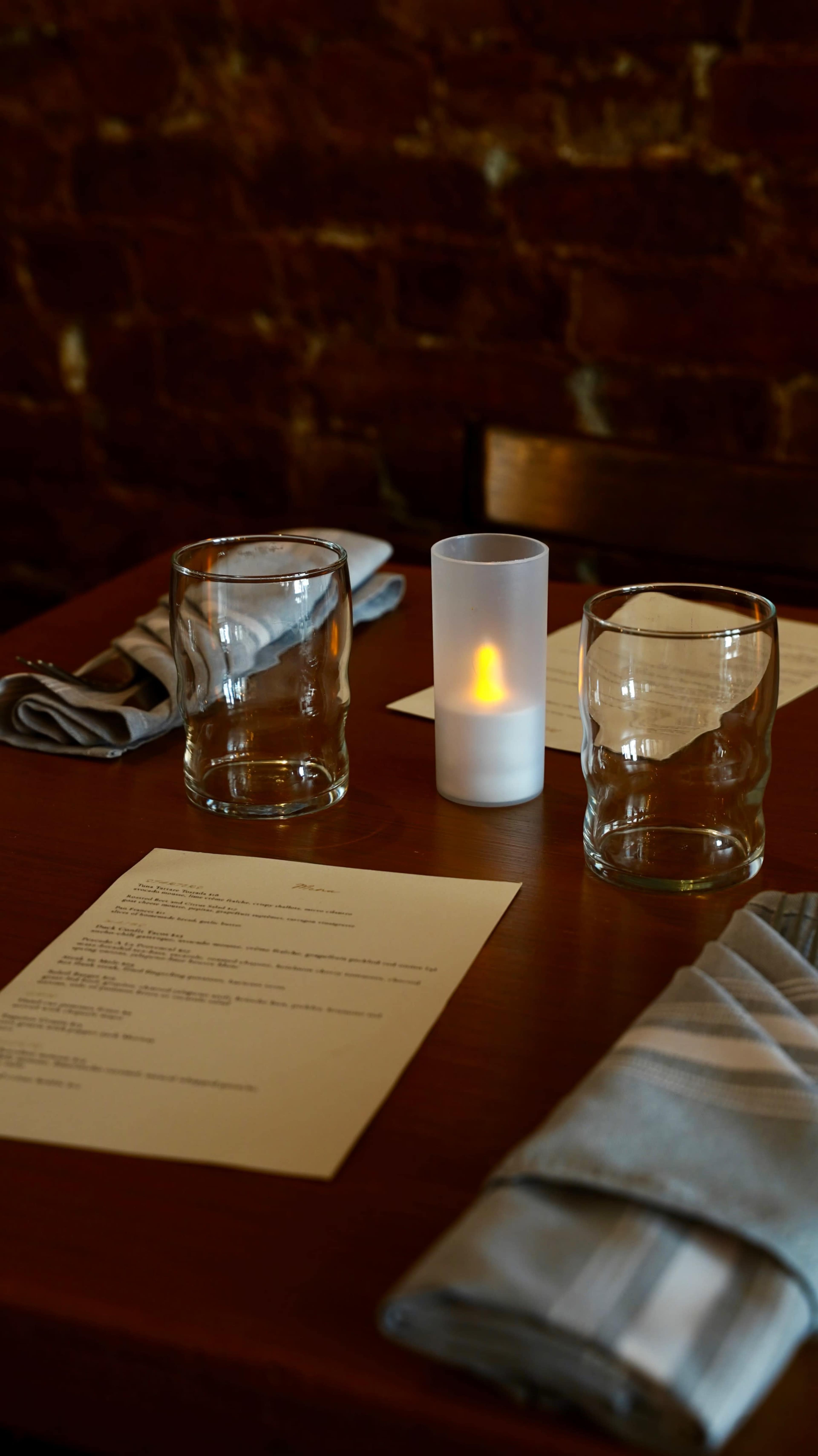 A wooden table is set with two empty glasses, a battery-operated candle, folded napkins, and a menu.
