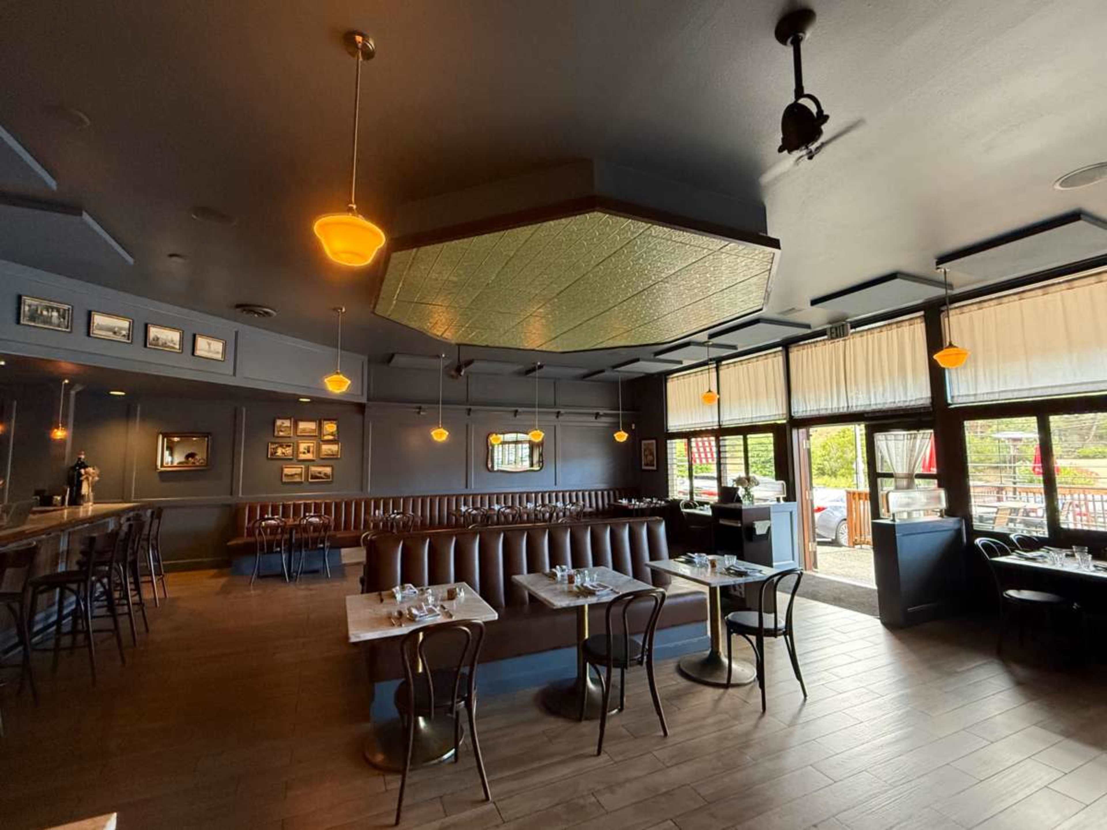 The interior of a restaurant features wooden flooring, brown leather banquettes, wooden tables, and large windows overlooking an outdoor patio.