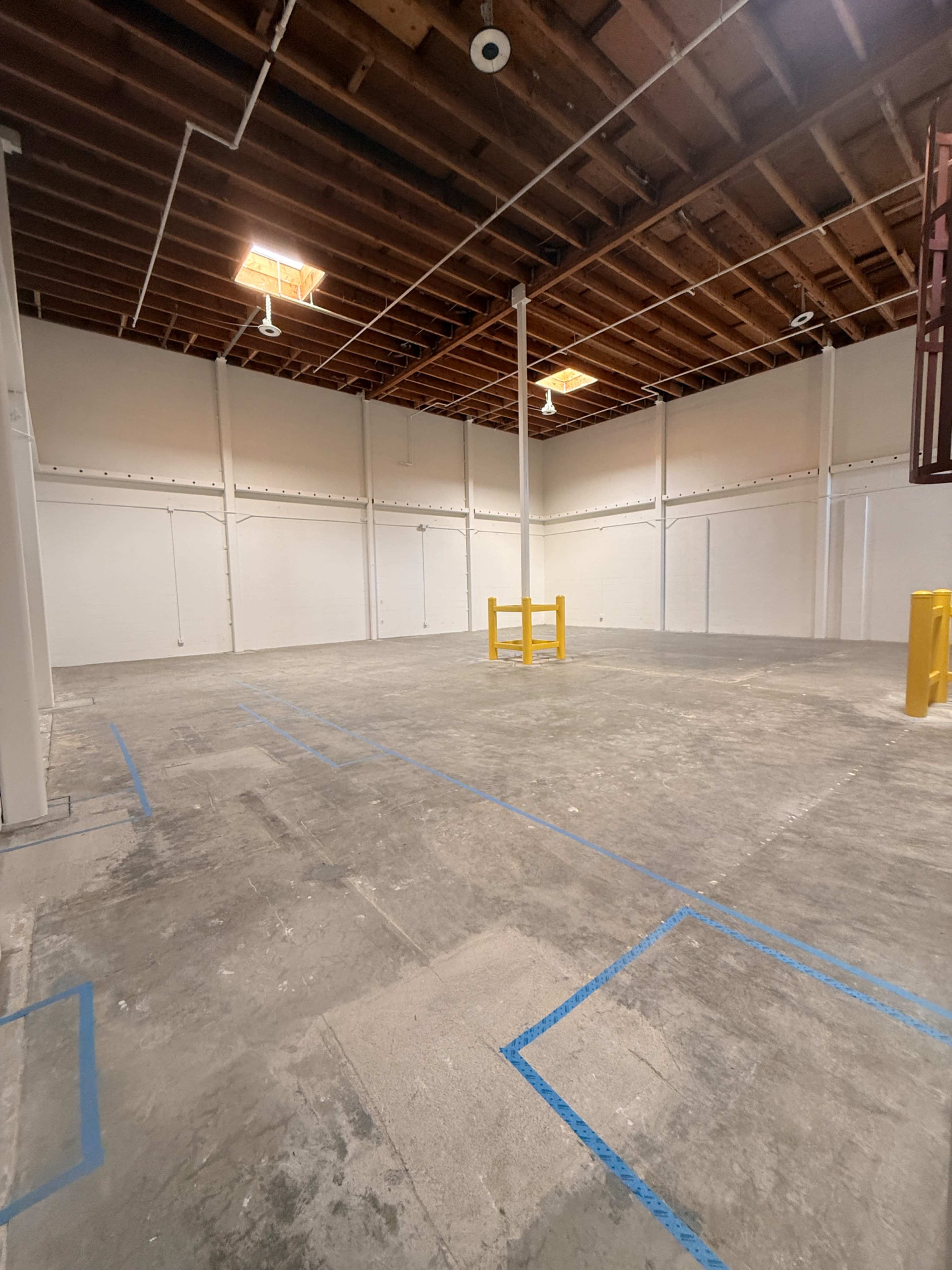 The image shows an empty interior space with bare concrete floors, wooden beams on the ceiling, and marked areas on the ground.