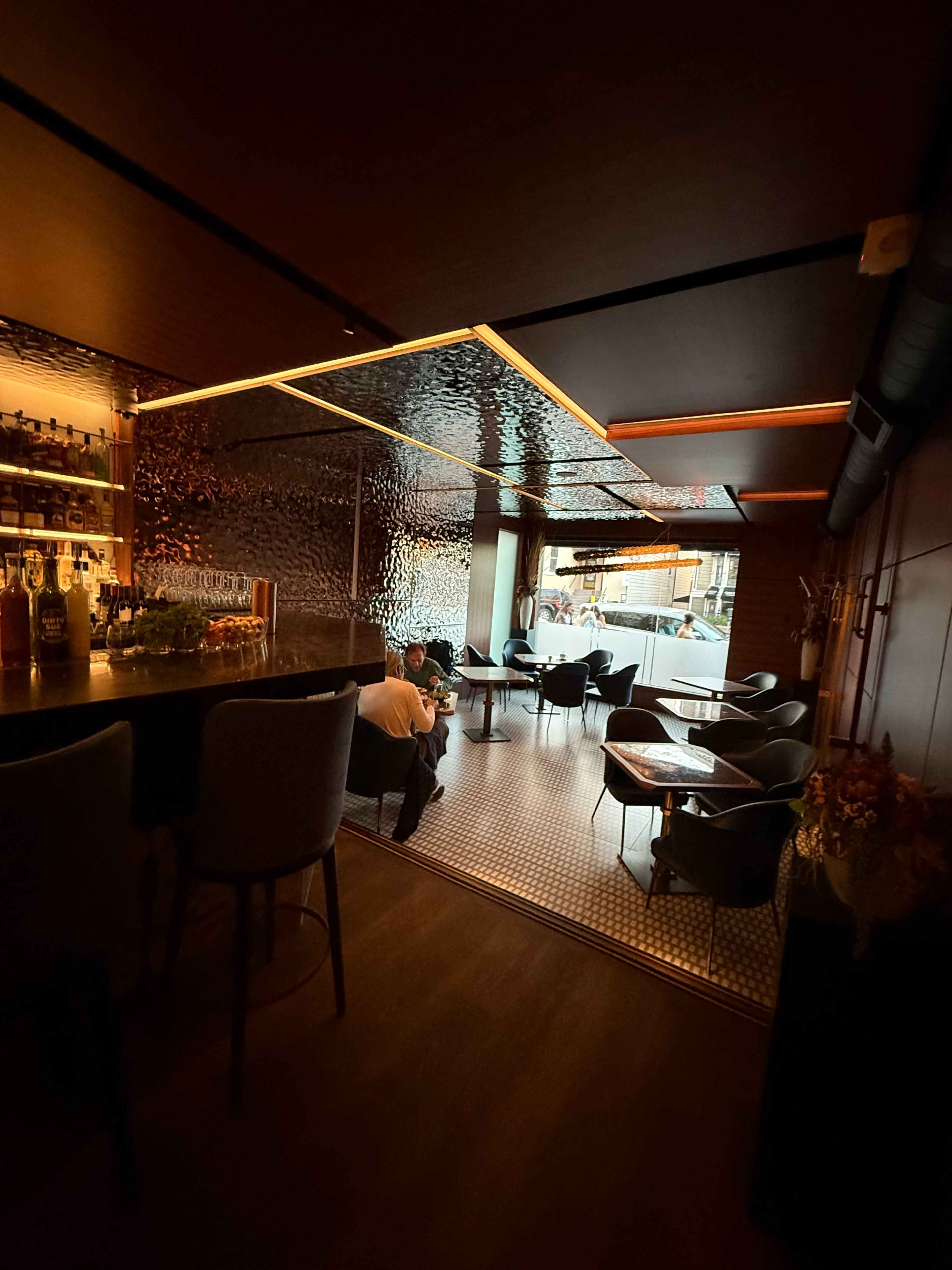 The image shows a dimly lit bar with a backlit counter, plush seating, and a view of the outside through large windows.