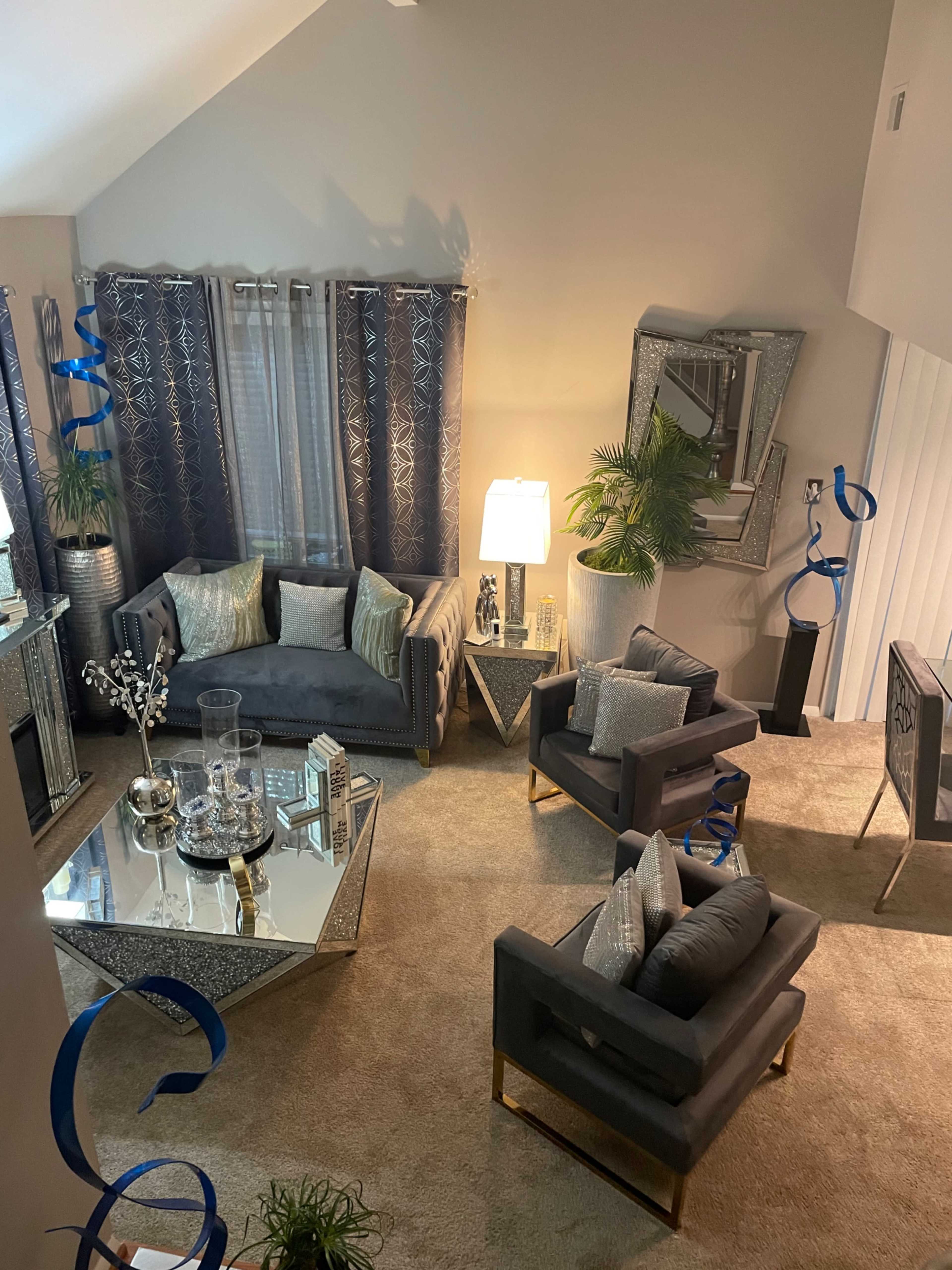 A modern living room furnished with a gray sofa, two matching chairs, a mirrored coffee table, and decorative plants, all arranged on a light-colored carpet.