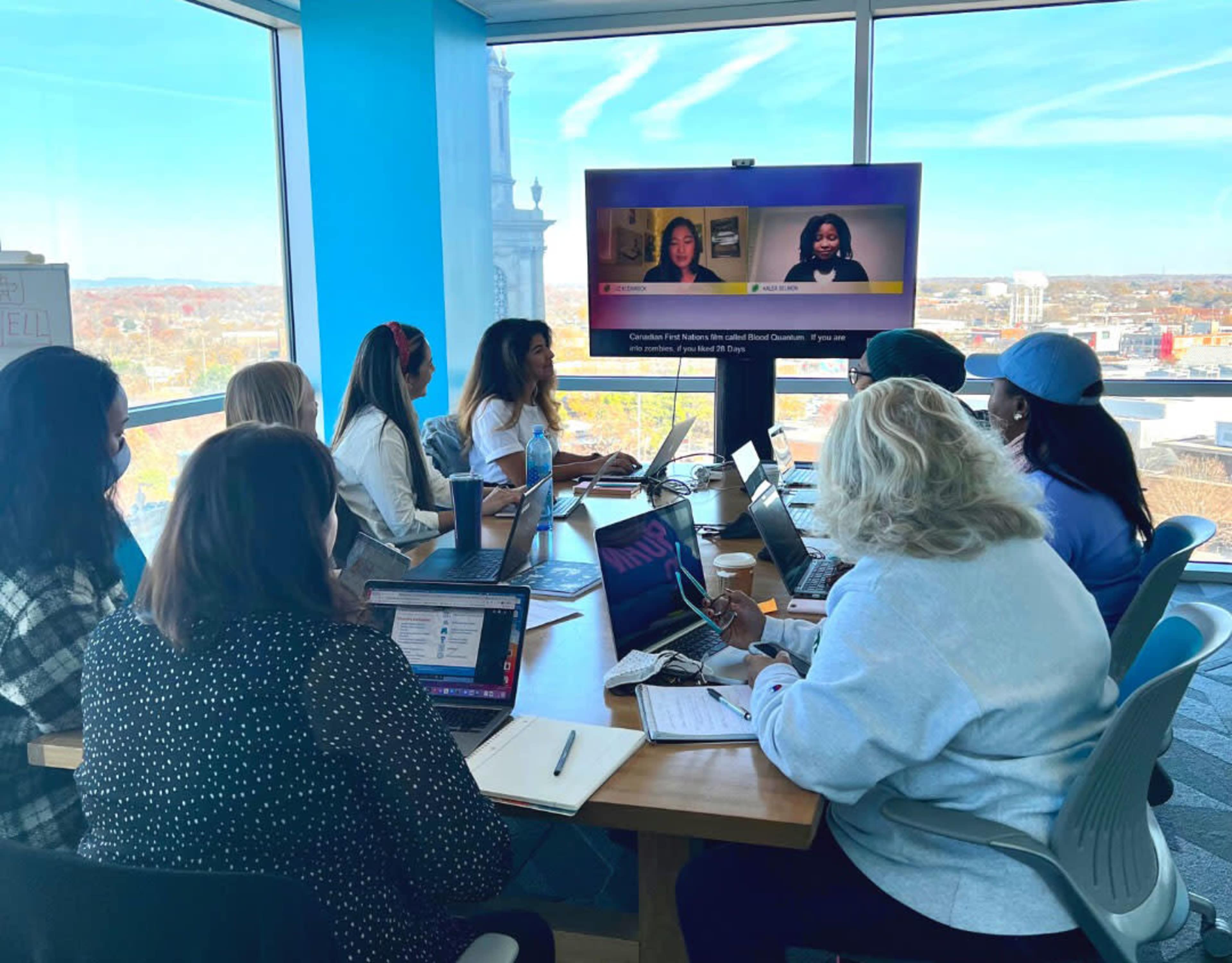 A group of eight people is sitting around a conference table with laptops, watching a presentation on a screen in a bright office setting.