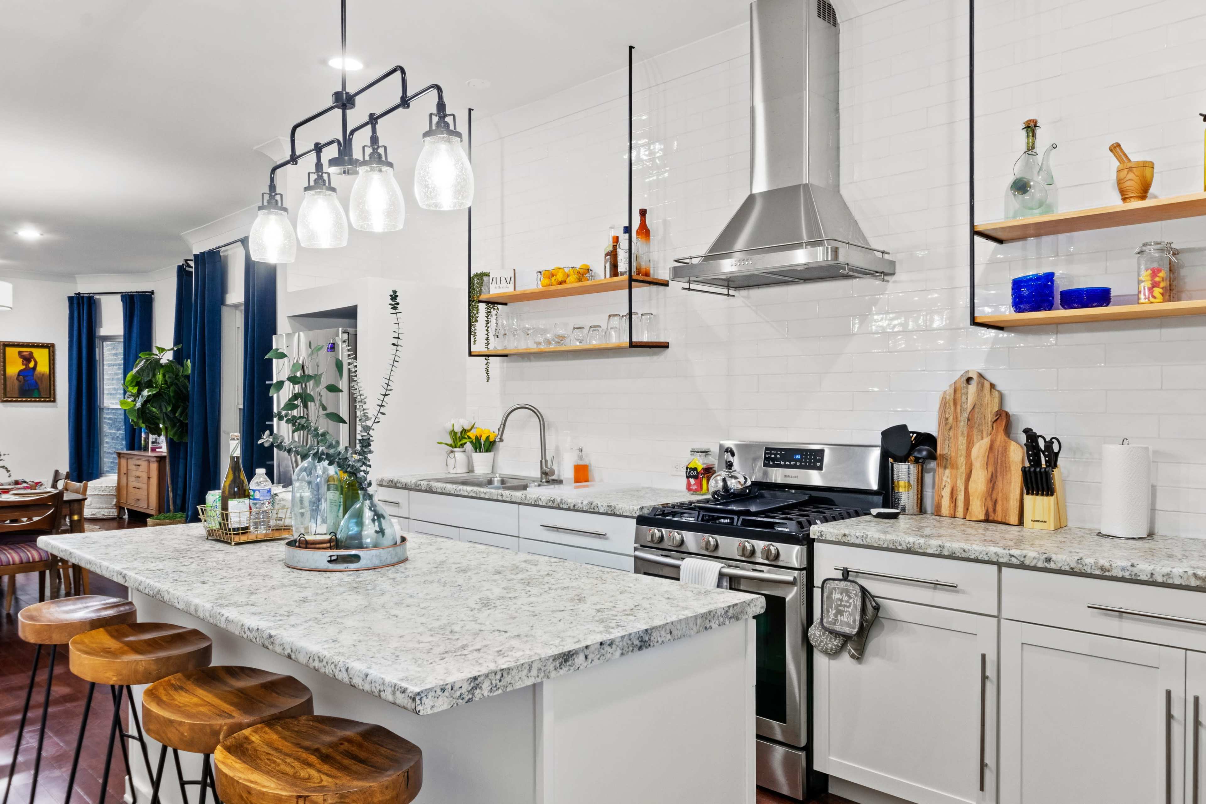 A modern kitchen features a large island with three wooden stools, sleek appliances, and open shelves displaying various kitchenware.