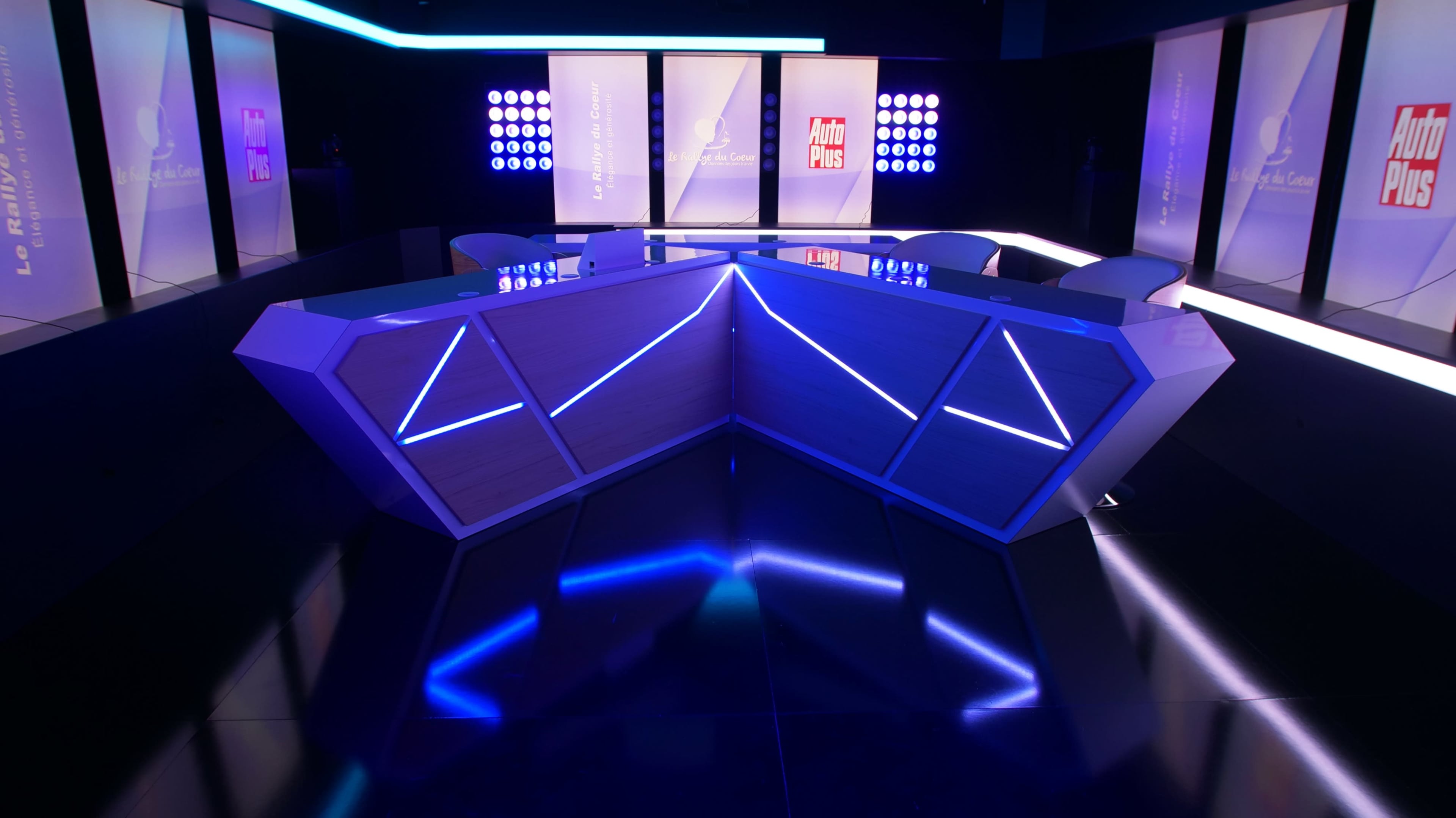 The image shows a modern studio set with a geometric table featuring blue lighting, surrounded by chairs and illuminated backdrop screens.