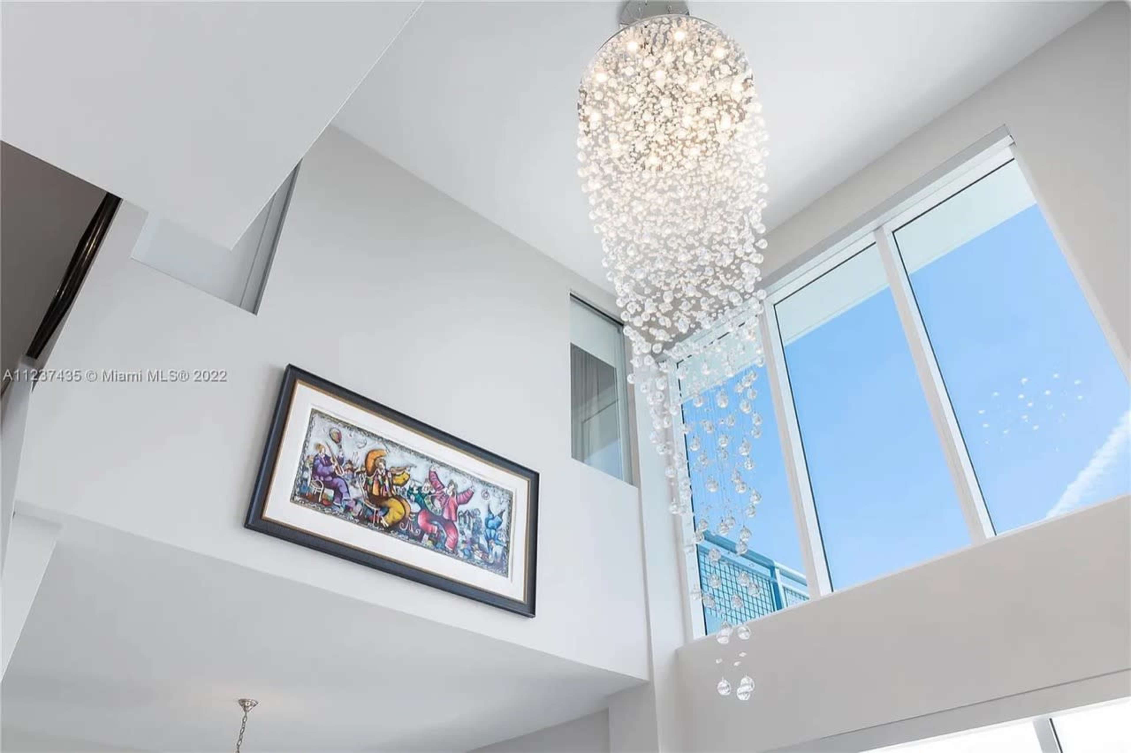 A modern interior features a large chandelier made of hanging beads above a colorful framed artwork, with large windows allowing natural light.
