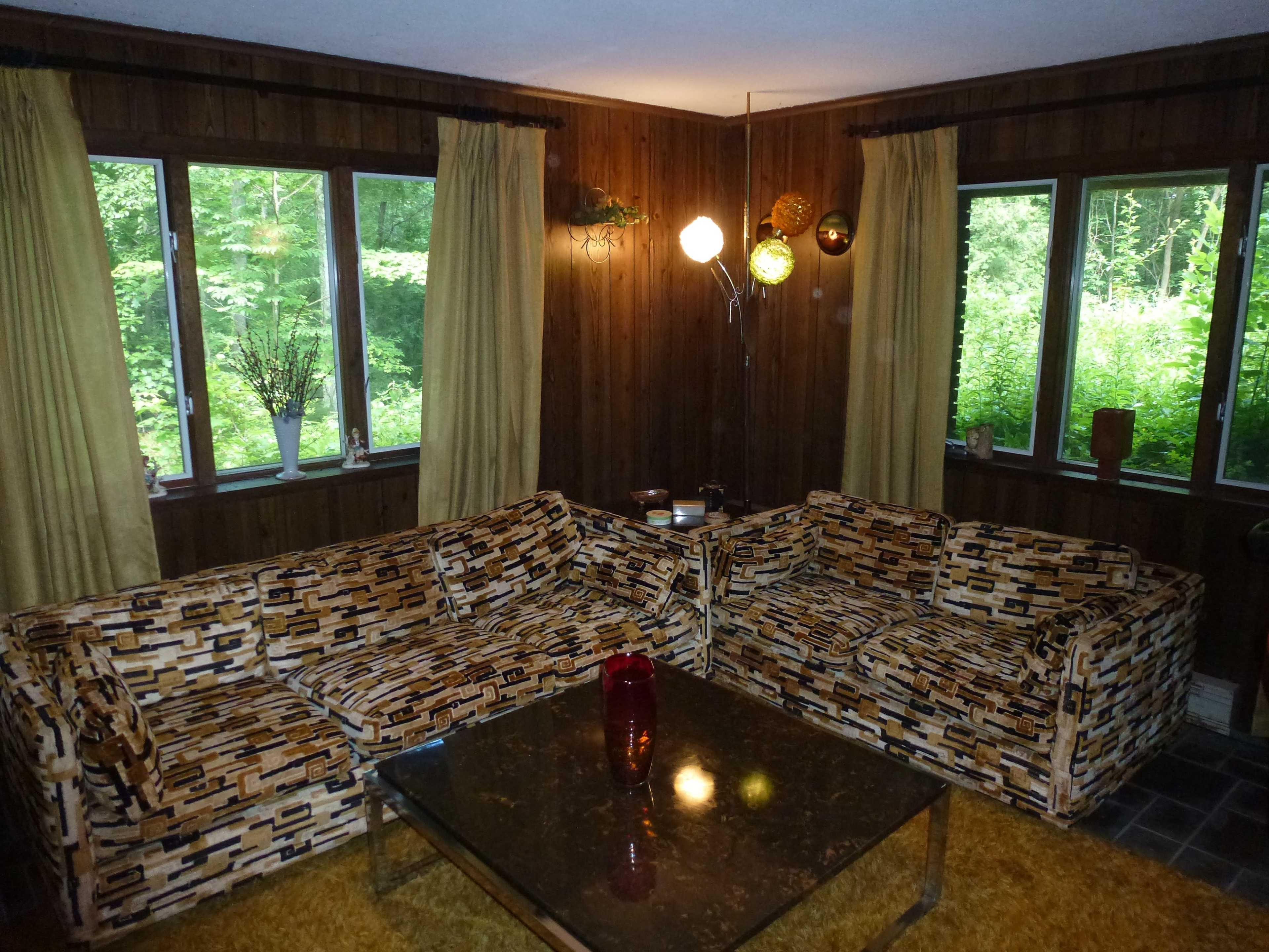 The scene features a living room with two patterned sofas, a glass coffee table, and large windows that overlook a green outdoor setting.