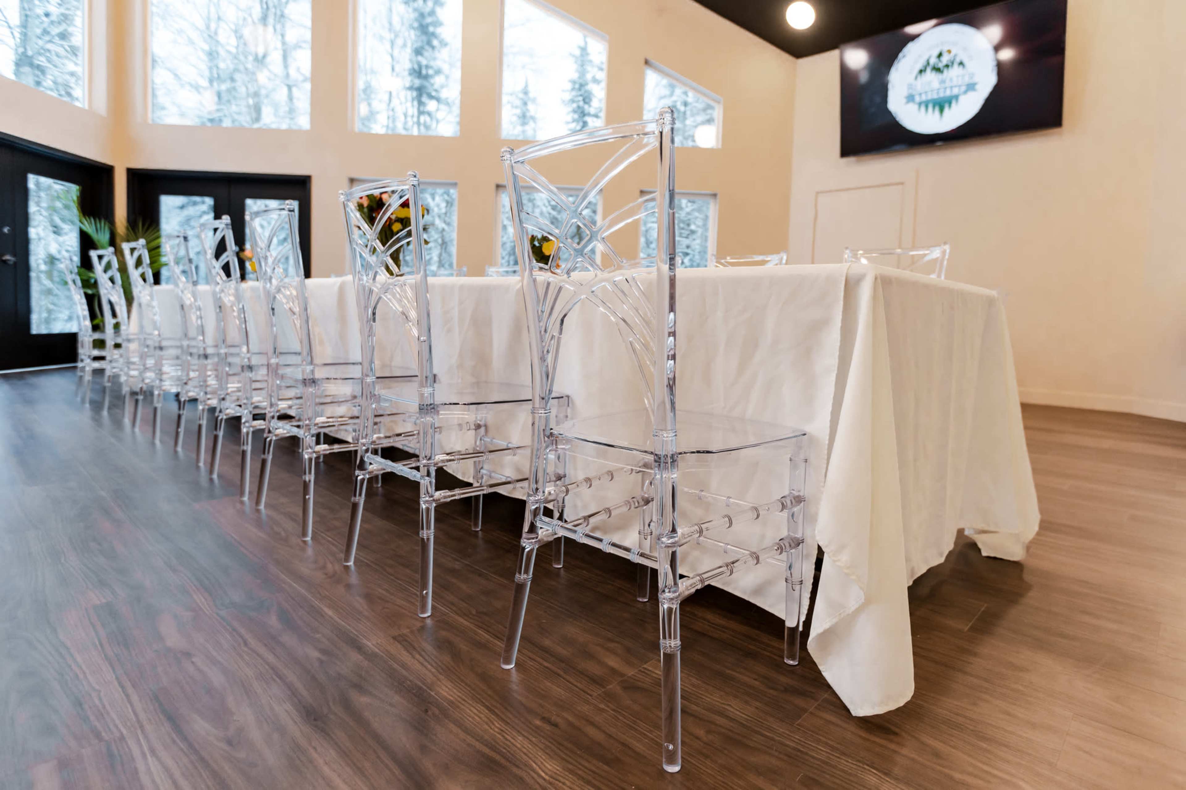 A long table dressed in a white tablecloth is set with transparent chairs in a room with large windows displaying a snowy landscape outside.