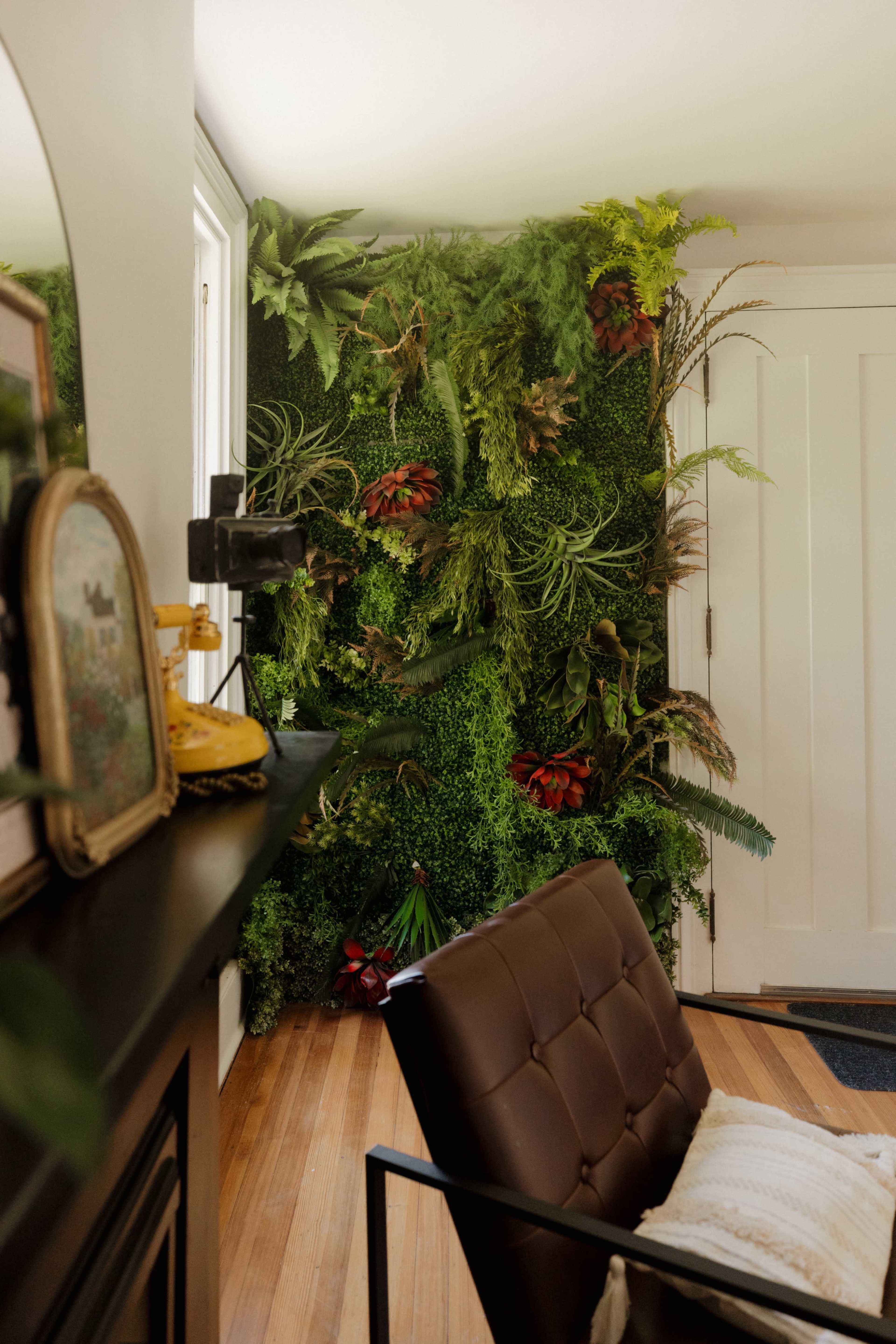The image shows a corner of a room featuring a vertical garden wall adorned with various green plants, framed by a modern chair and a decorative mirror on a black console table.