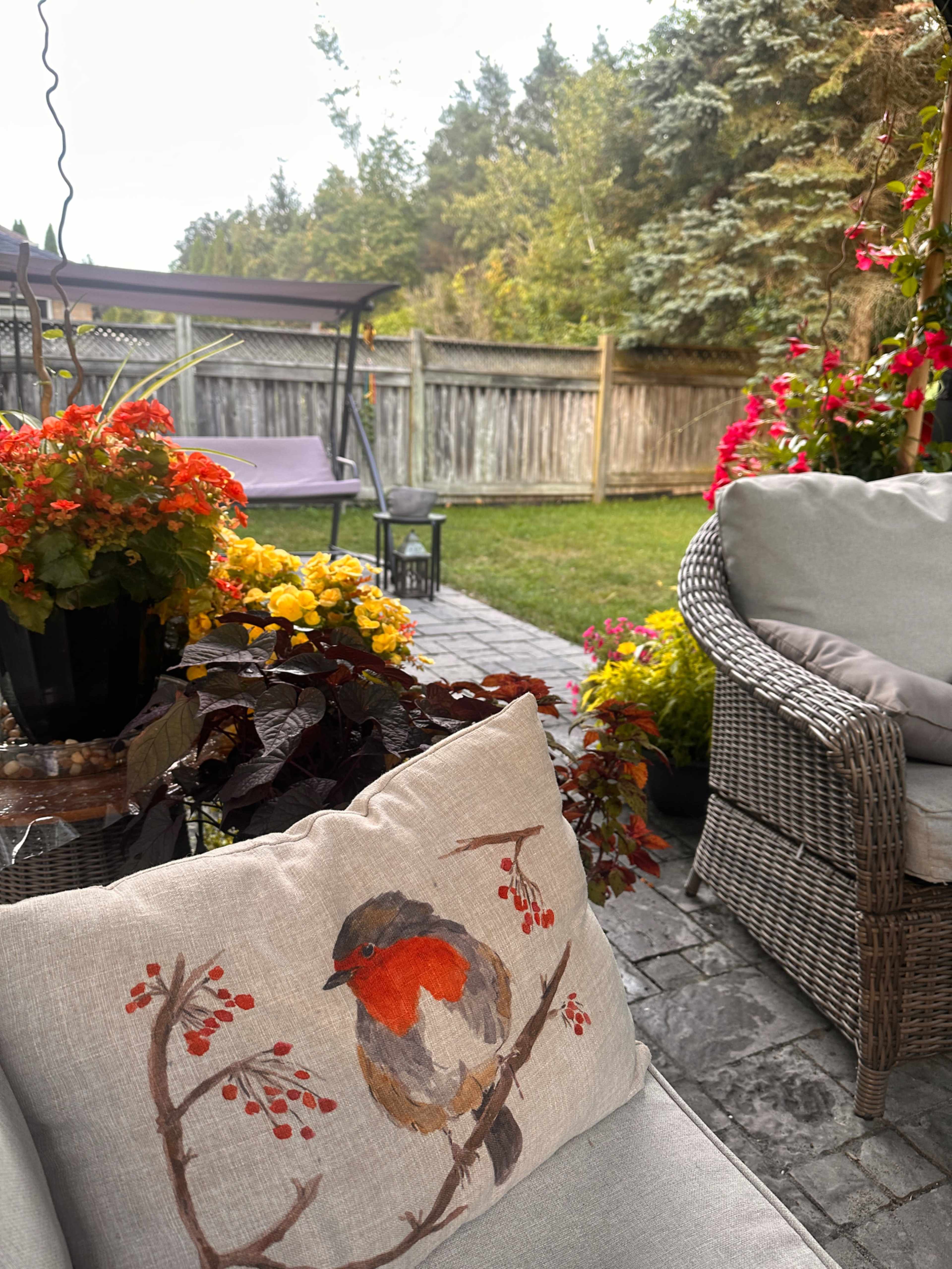 A cozy outdoor seating area features a decorative pillow with a bird illustration, surrounded by colorful flowers and greenery.