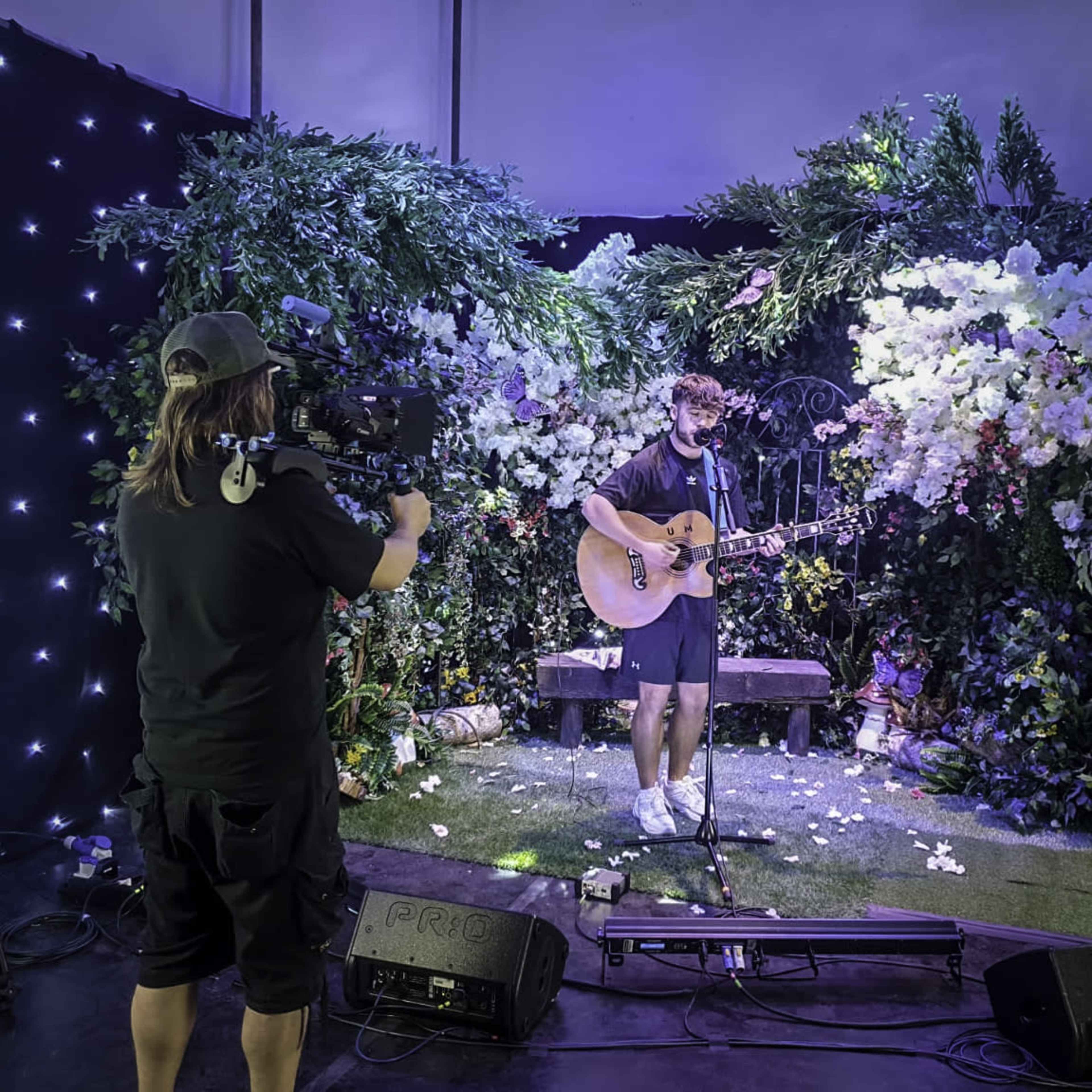 A musician performs on stage with an acoustic guitar in front of a floral backdrop while being filmed by a cameraman.