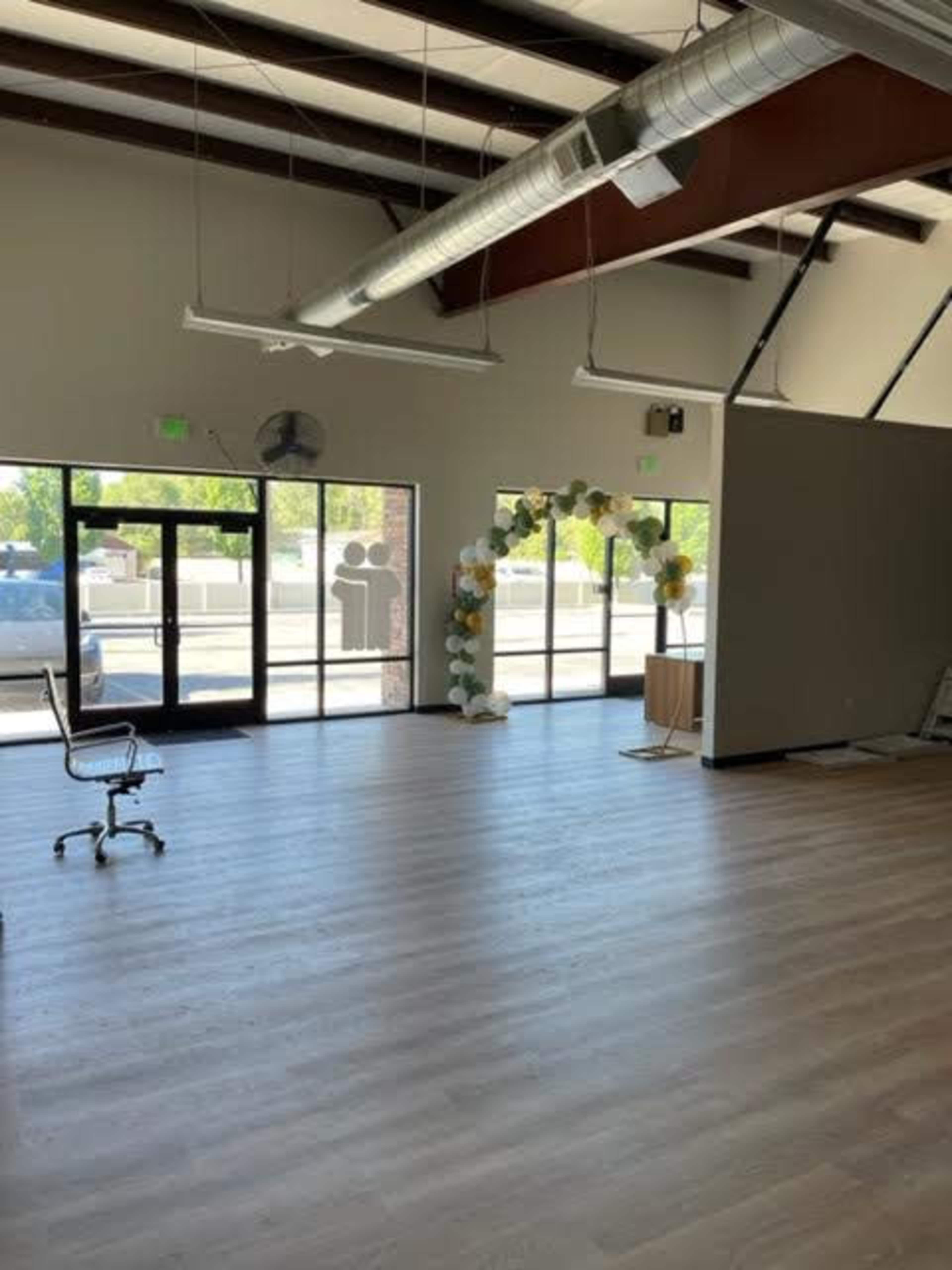 A spacious, empty room with wooden flooring and large windows, featuring an arch decorated with flowers near a table.