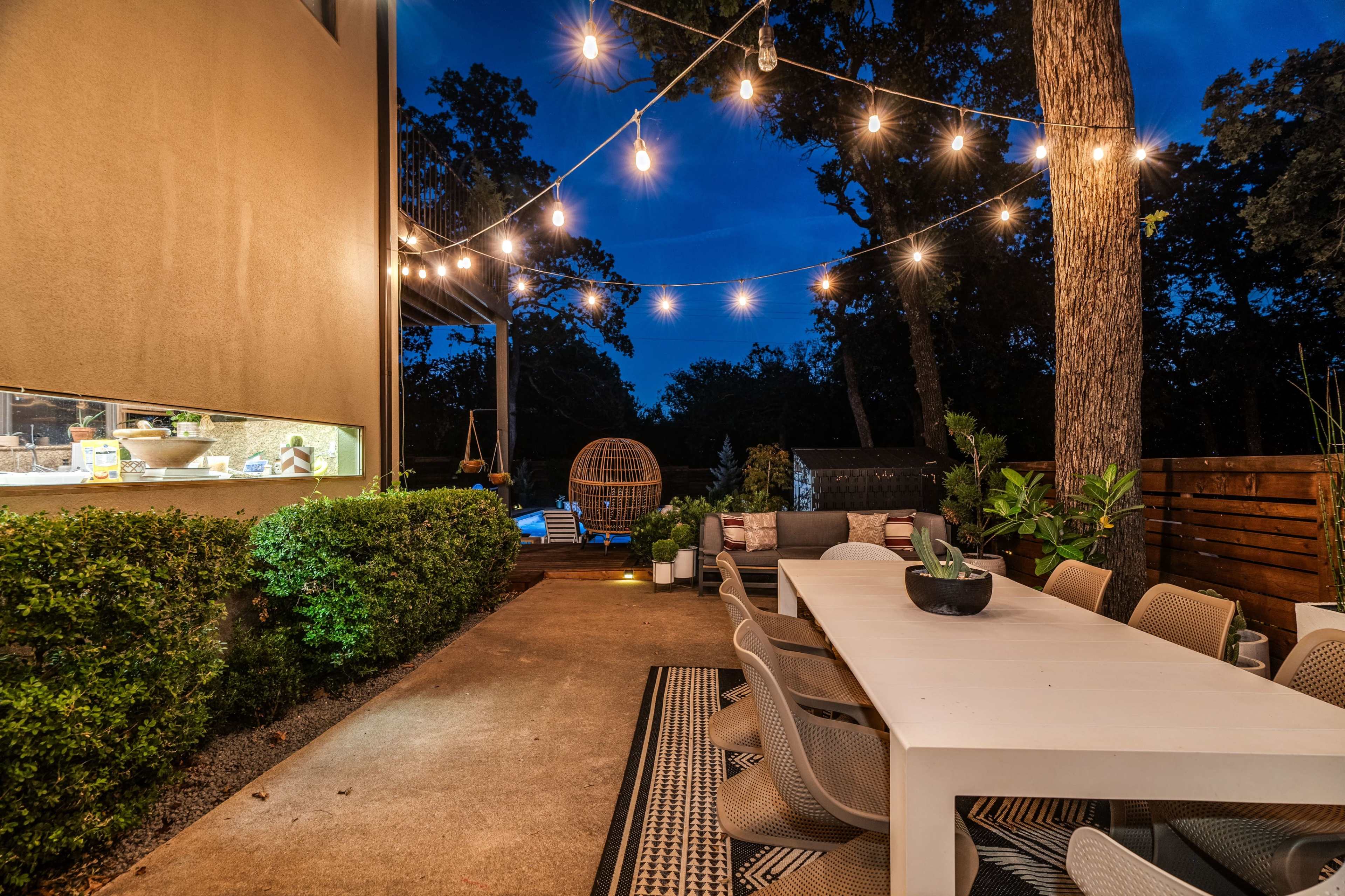 A modern outdoor patio featuring a long dining table, string lights overhead, and greenery surrounding the space.