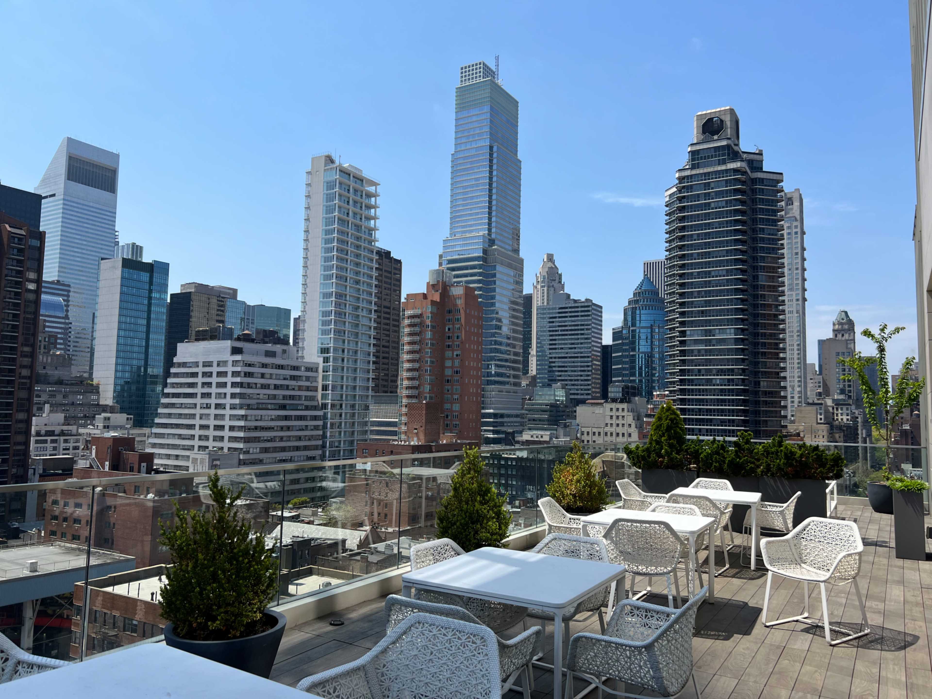 A rooftop terrace with white seating and greenery, overlooking a skyline of skyscrapers.