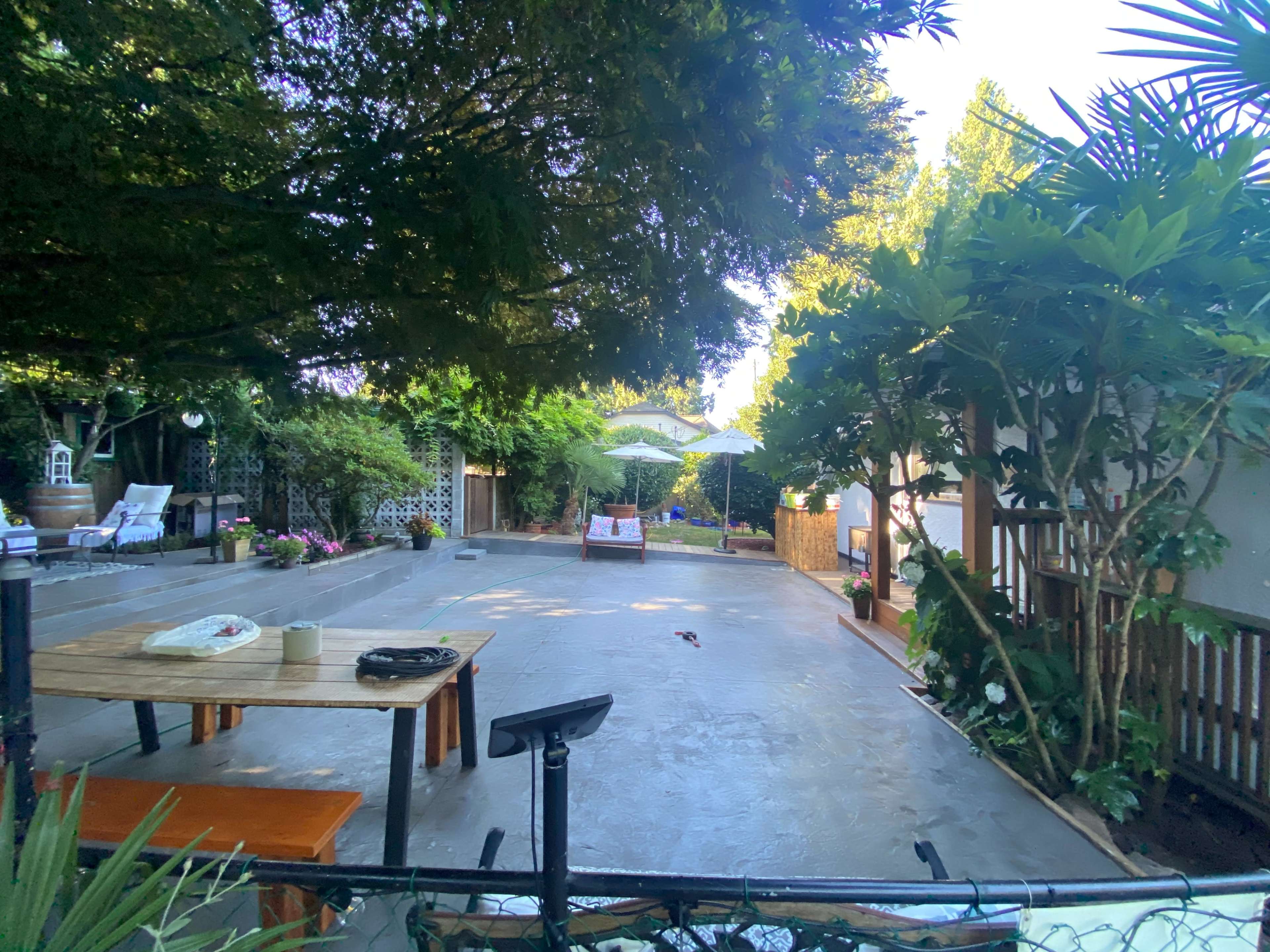 A spacious outdoor area featuring a concrete floor, several plants, a table with items on it, and lounge chairs under an umbrella.