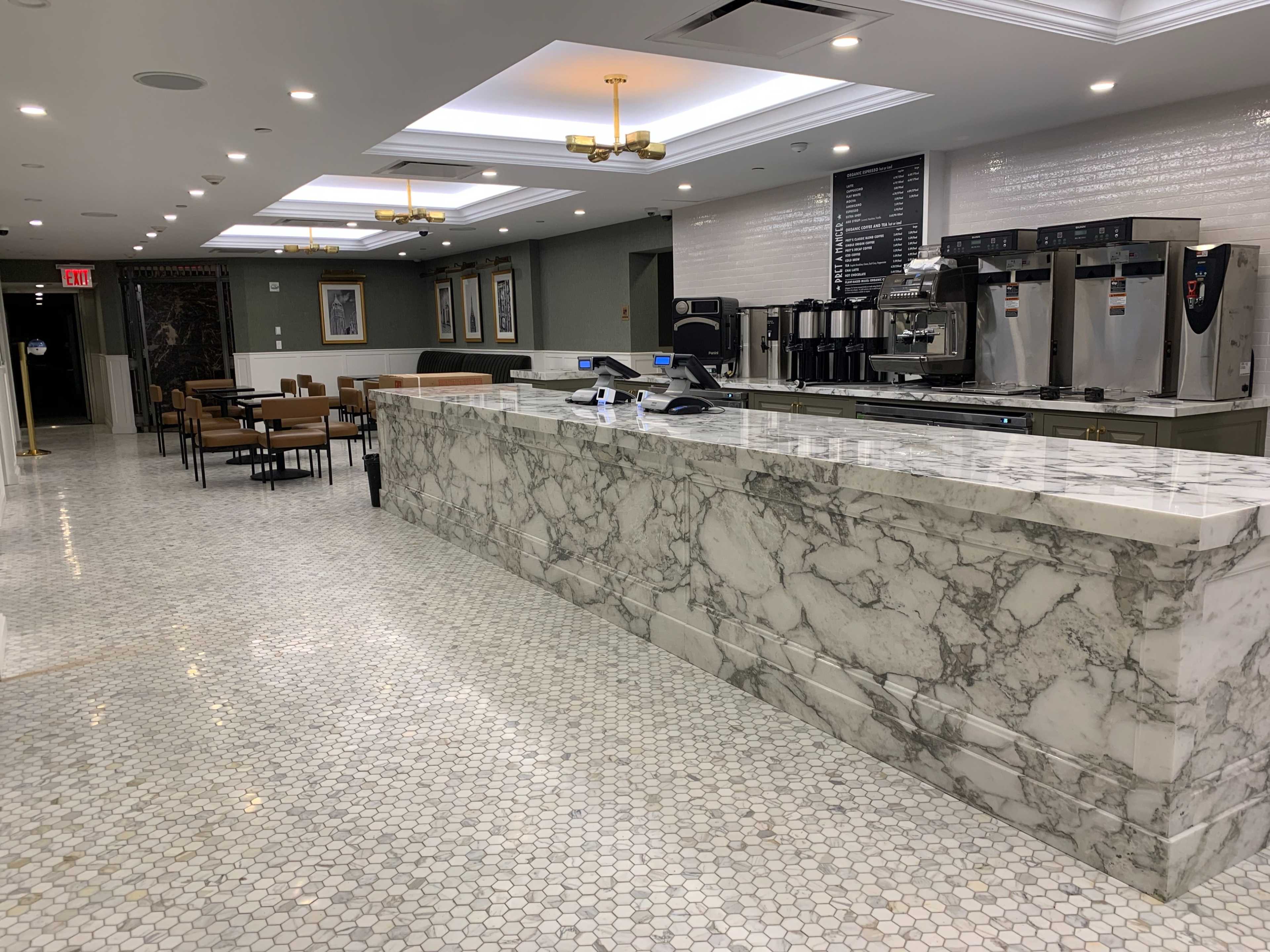 The image shows a modern cafe interior with a marble countertop, coffee machines, and seating arrangements, featuring hexagonal tiled flooring.