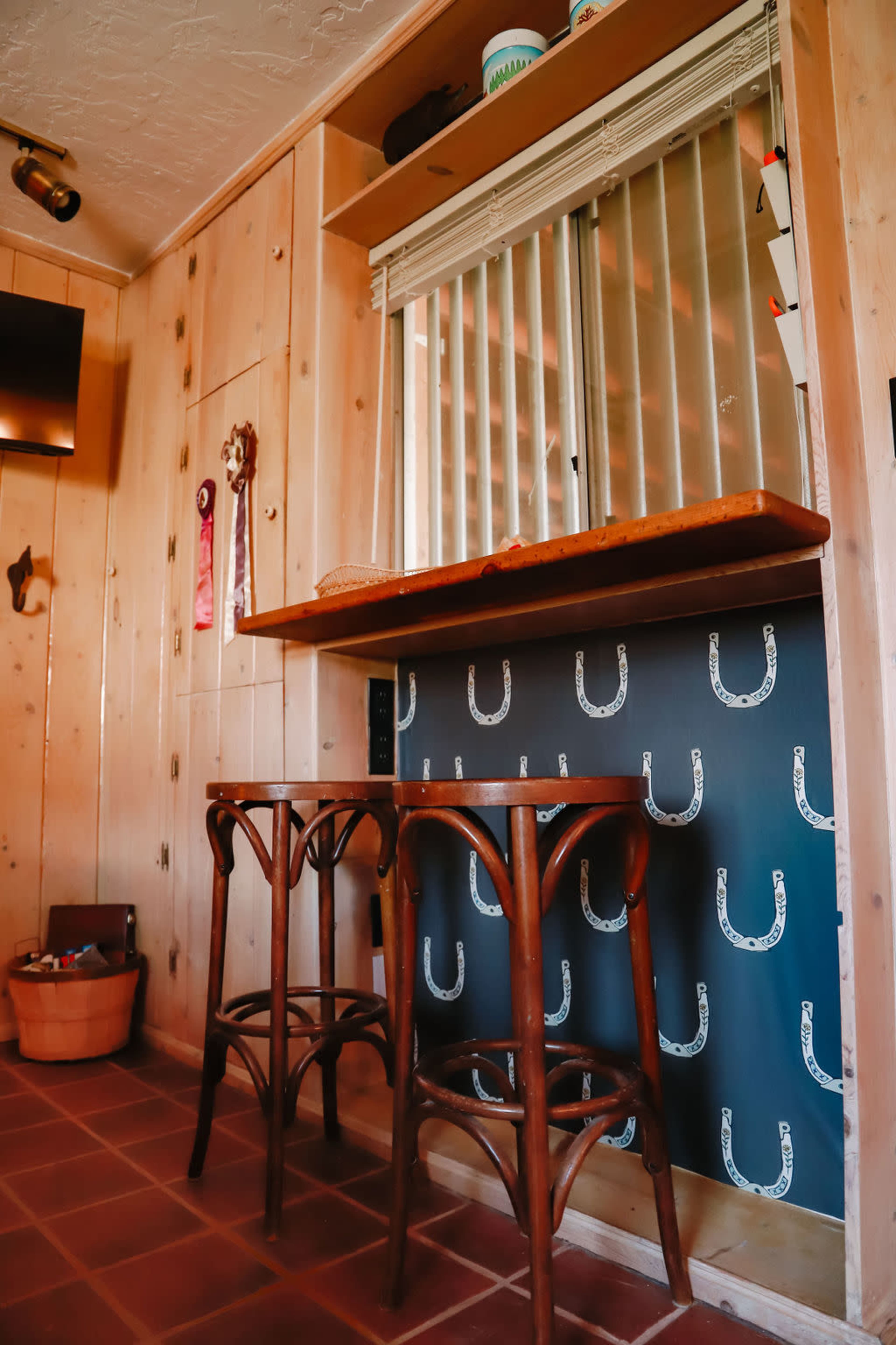 The image shows a wooden interior space featuring a bar area with two wooden stools and a wall decorated with horse shoe patterns.