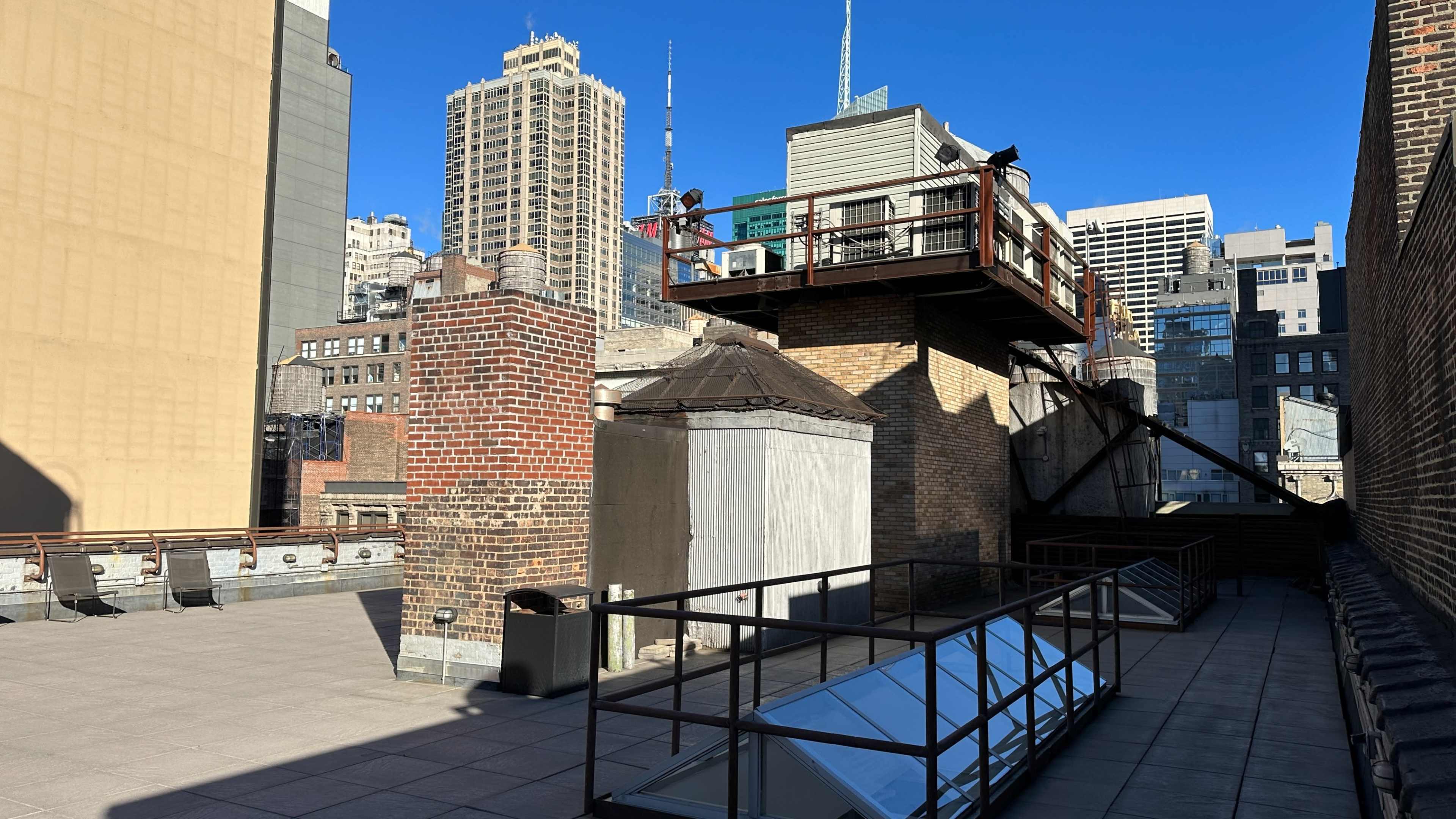 The image shows a rooftop area with a brick chimney, a gray storage structure, and a modern building with a patio overlooking a city skyline.