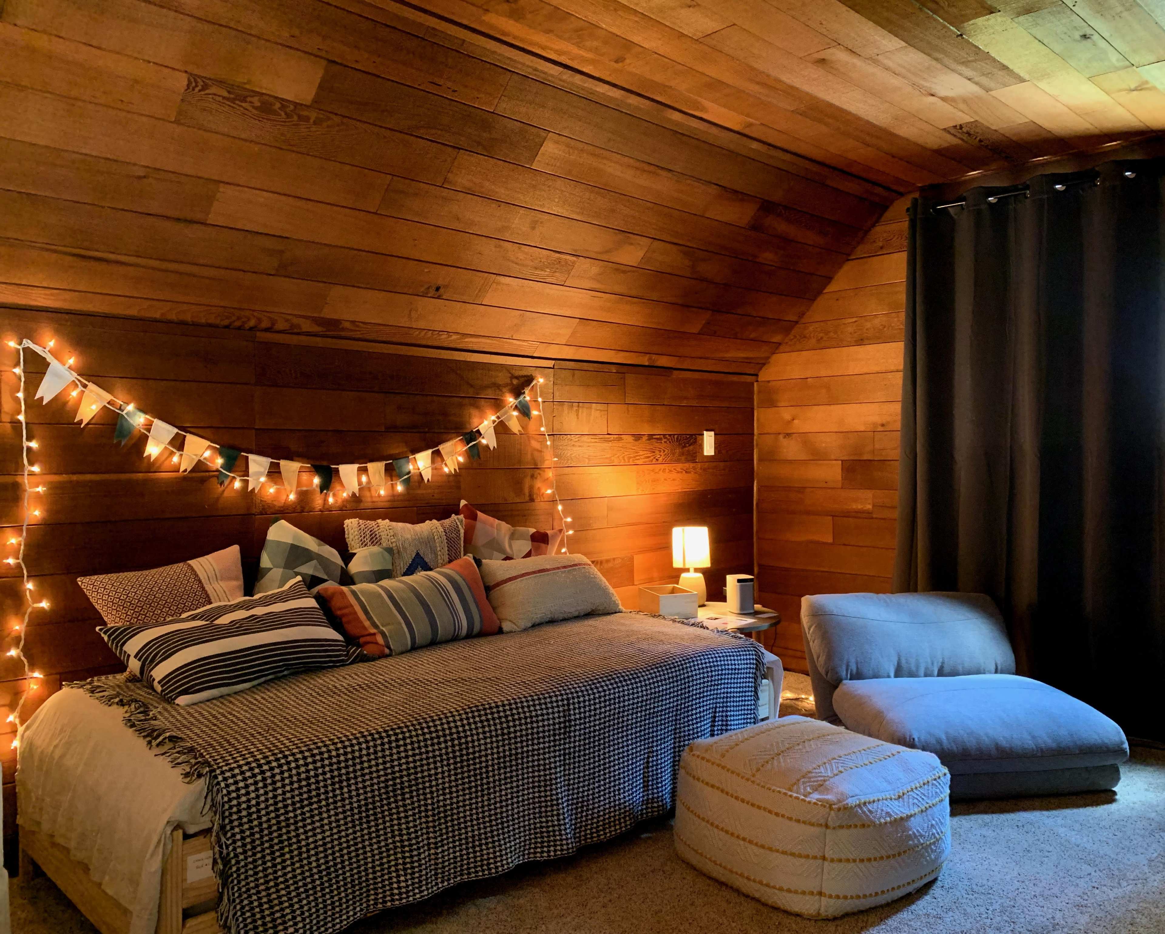 A cozy bedroom with a wooden ceiling, a bed adorned with various pillows, a small nightstand with a lamp, and a seating area with a large cushion and pouf.