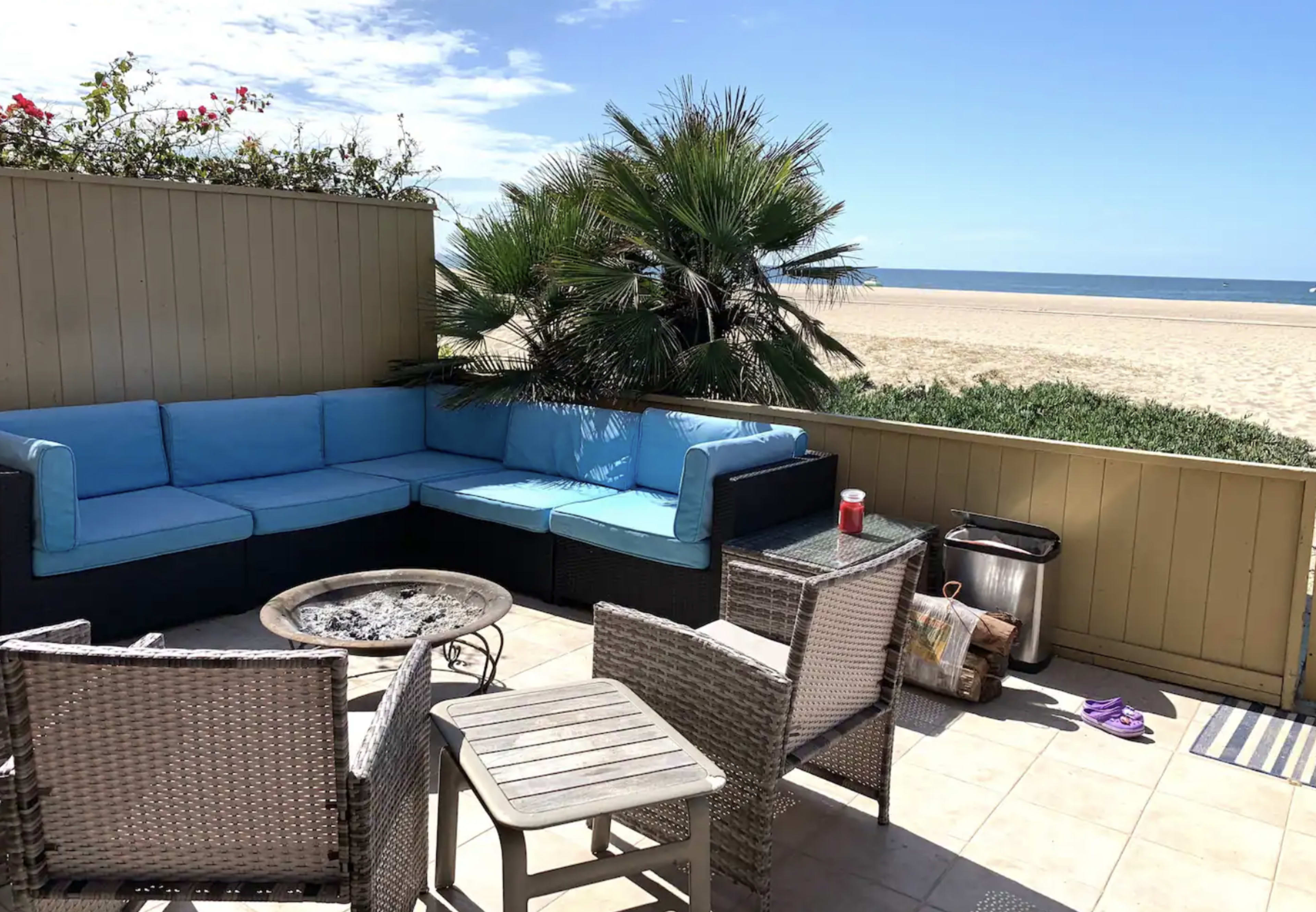 A patio features a blue sectional sofa, a fire pit, and a dining set with a view of the beach and ocean beyond.