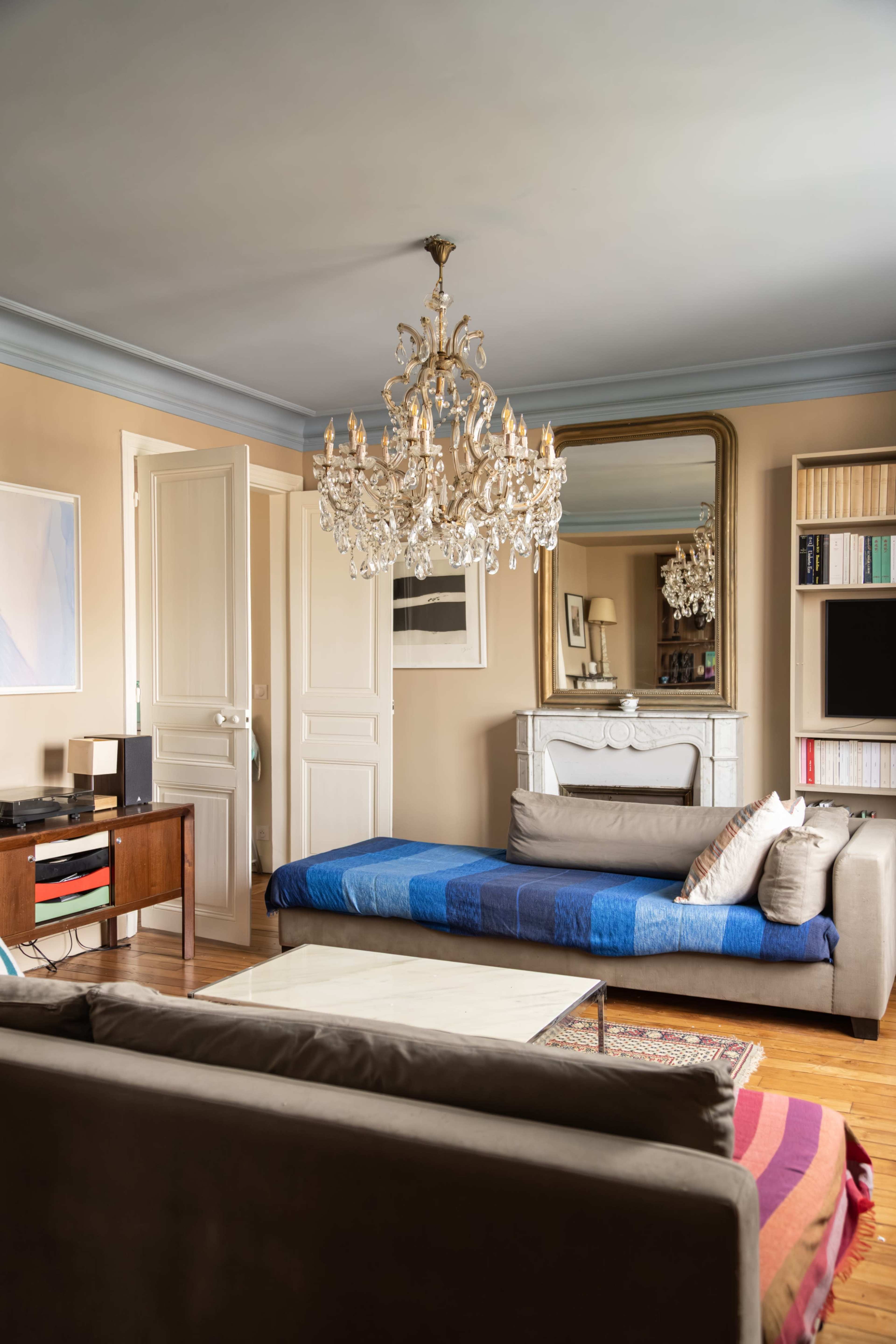 The image shows a stylish living room featuring a chandelier, a large mirror, a blue sofa, and a wooden coffee table.