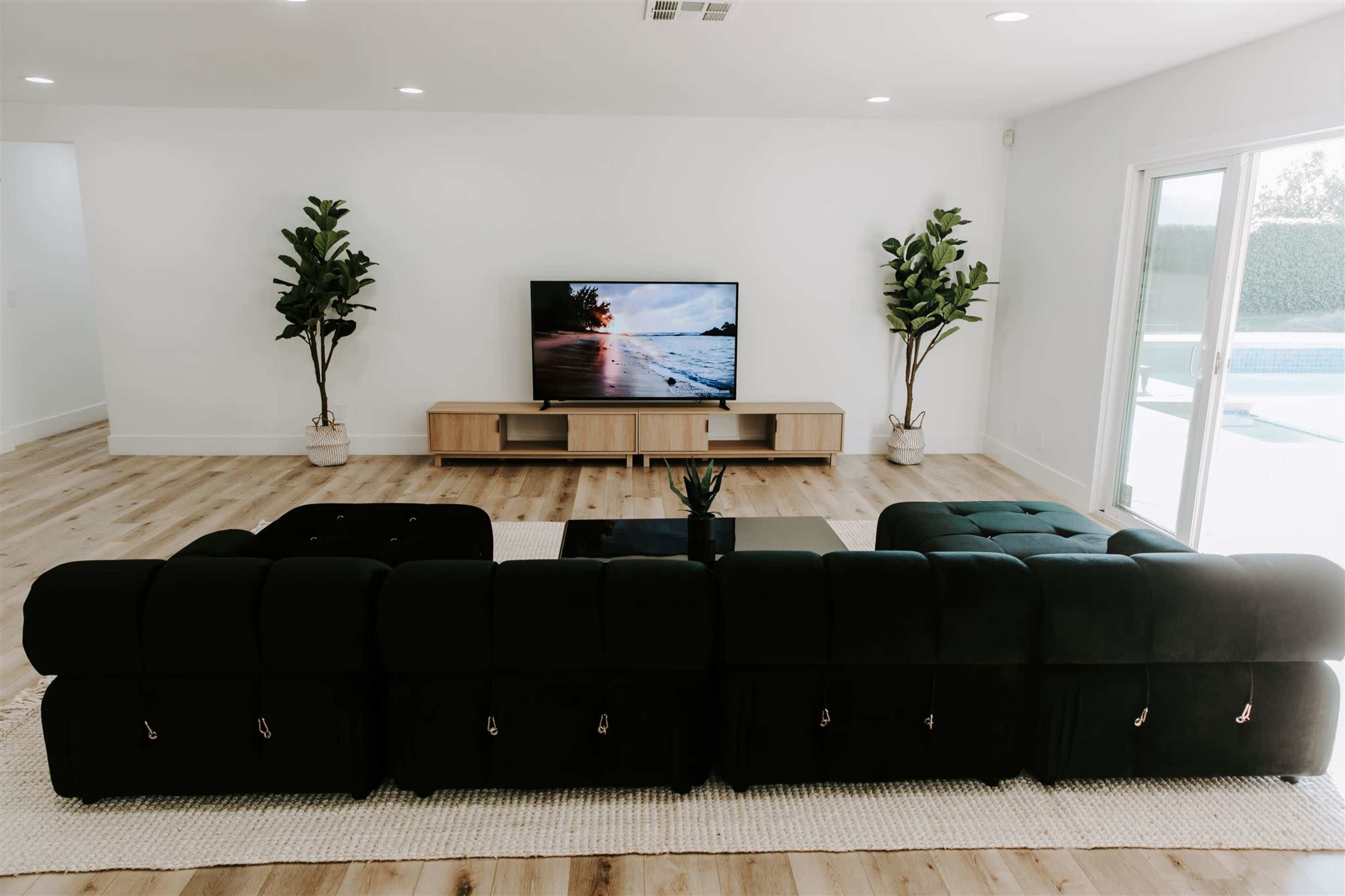 A modern living room features a black sectional sofa facing a television mounted on a light wood media console, with two potted plants flanking the sides.