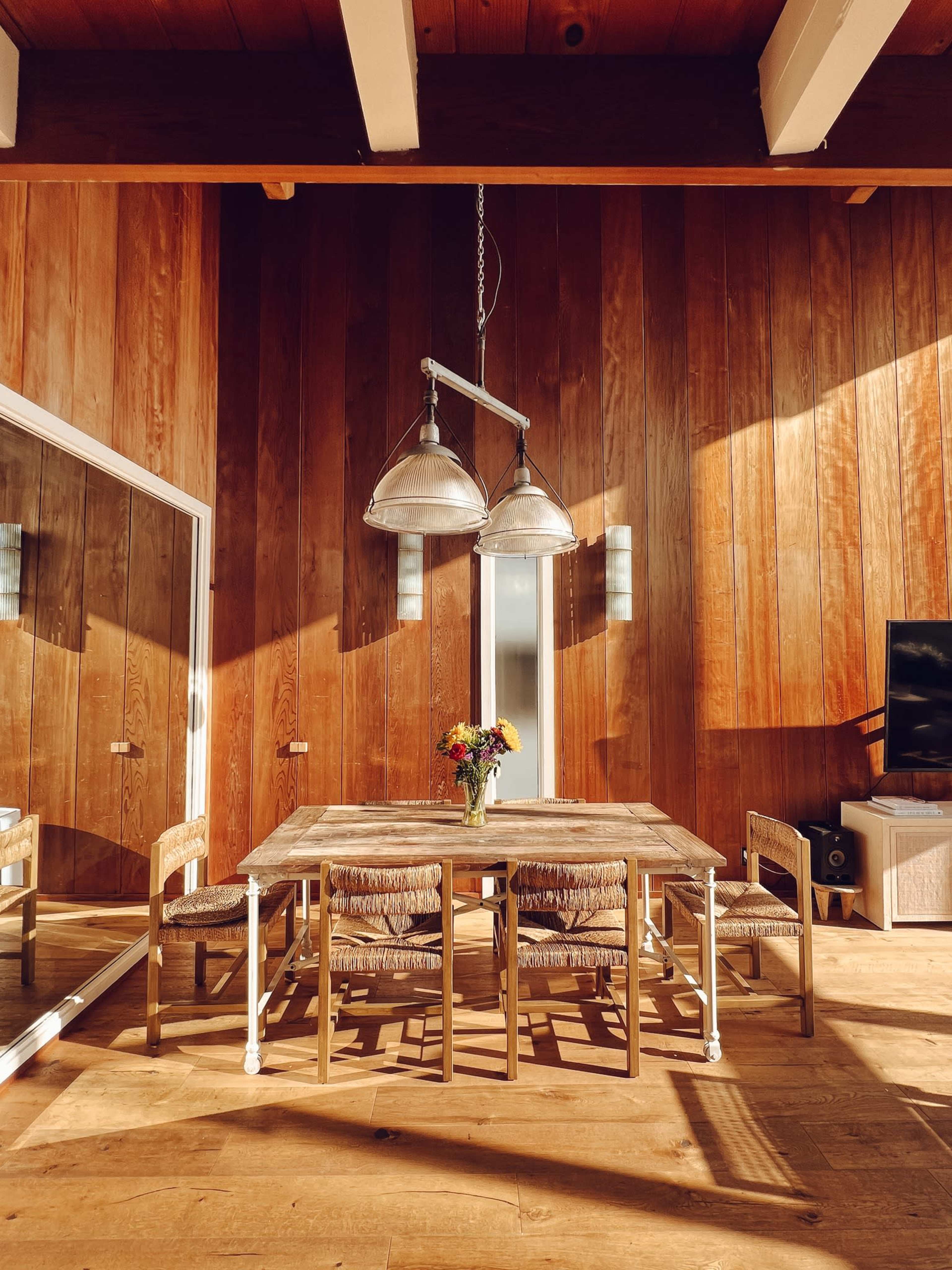 A dining area features a wooden table with chairs and a vase of flowers, illuminated by three pendant lights above.