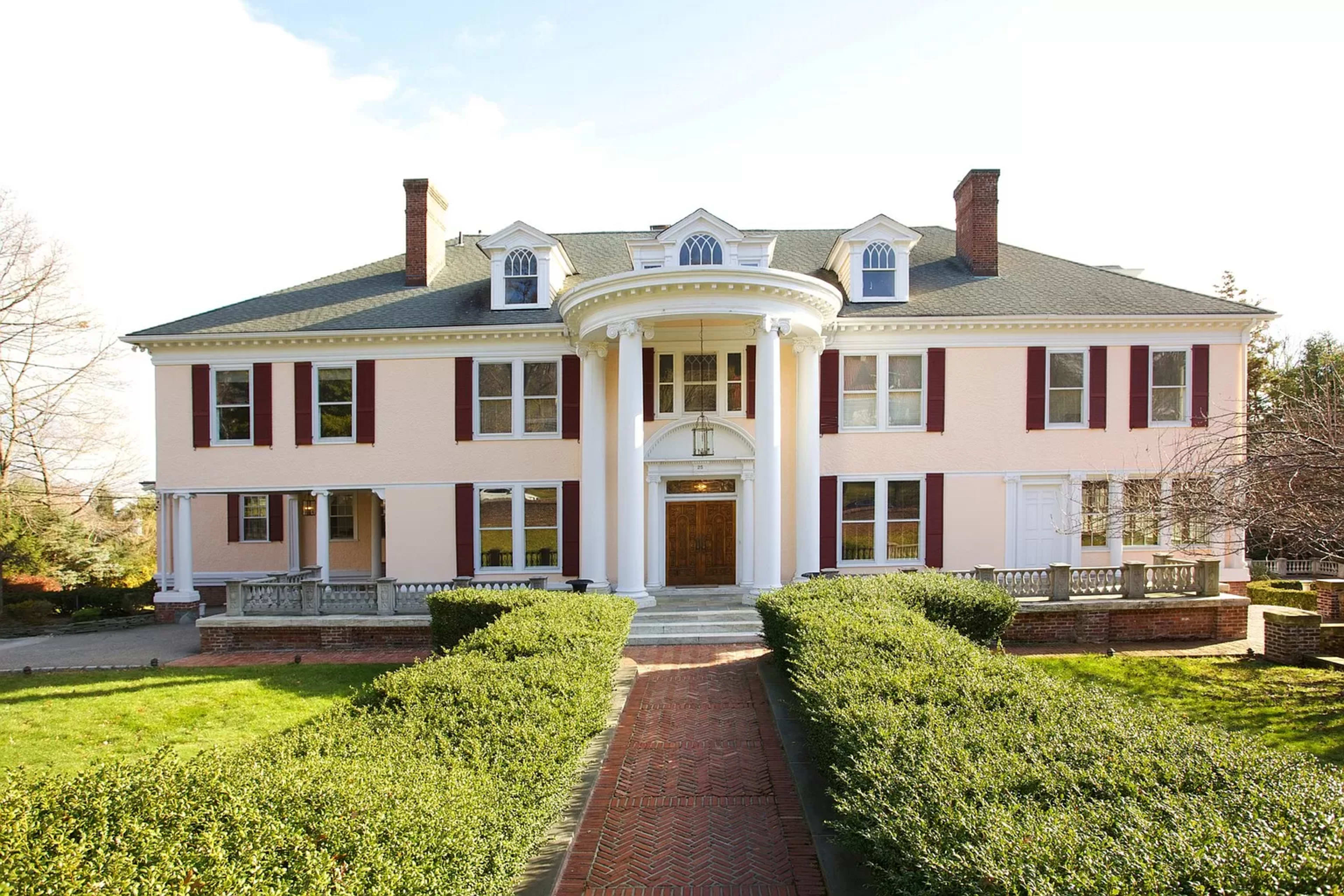 A large, two-story mansion with white columns and pink siding is framed by manicured hedges and a brick path leading to the front door.