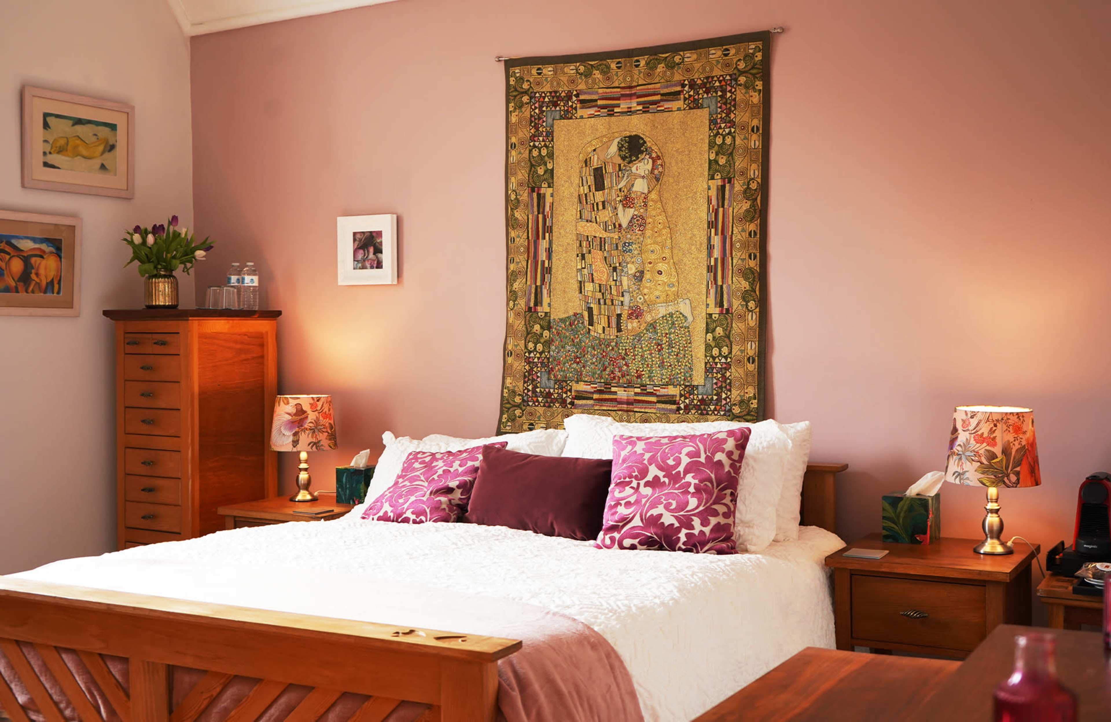 The image depicts a cozy bedroom with a large bed centered against a wall adorned with a decorative tapestry, flanked by nightstands with lamps.