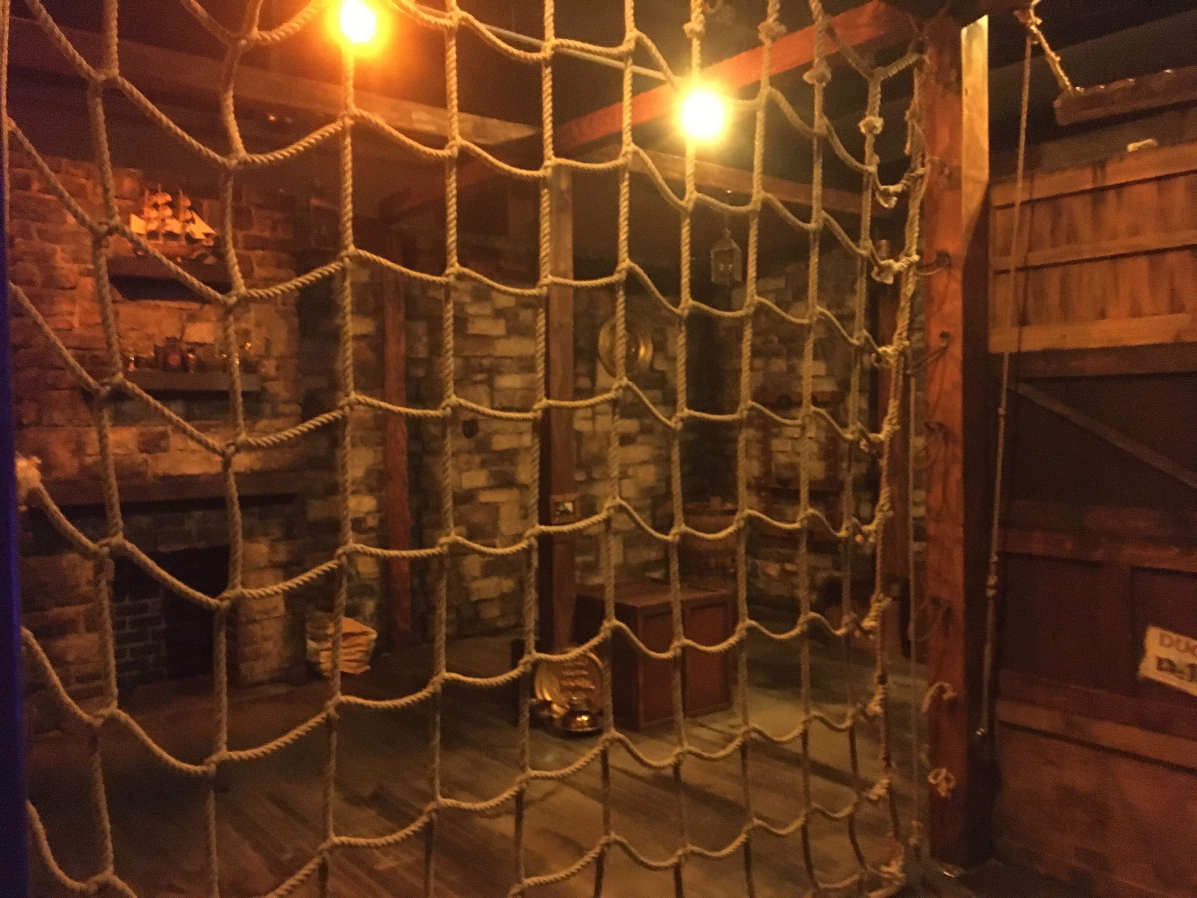 A dimly lit room with stone walls, wooden beams, and a large rope net hanging from the ceiling.