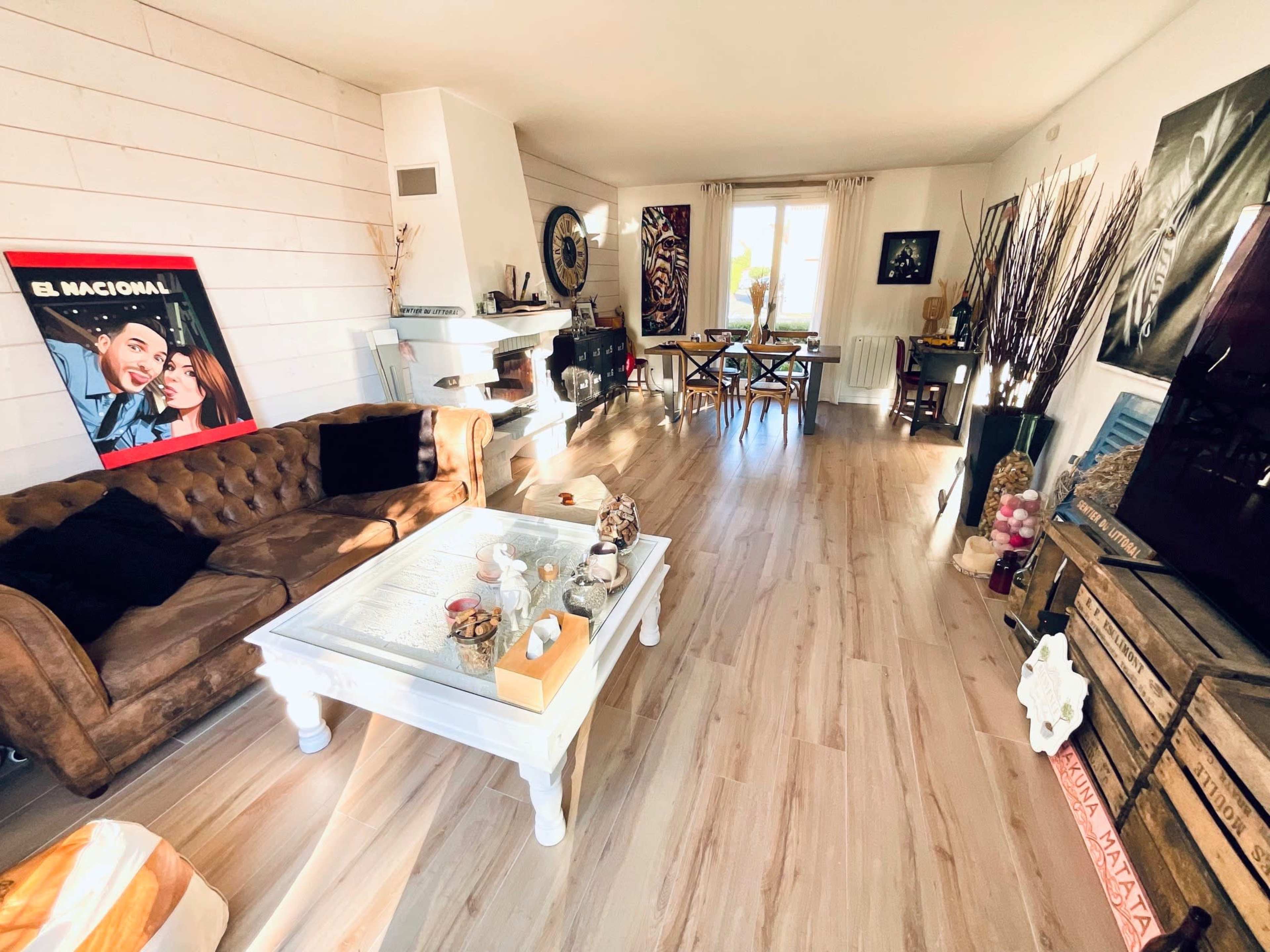 A spacious living room features a brown leather sofa, a wooden coffee table, and a fireplace, with large windows allowing natural light in.