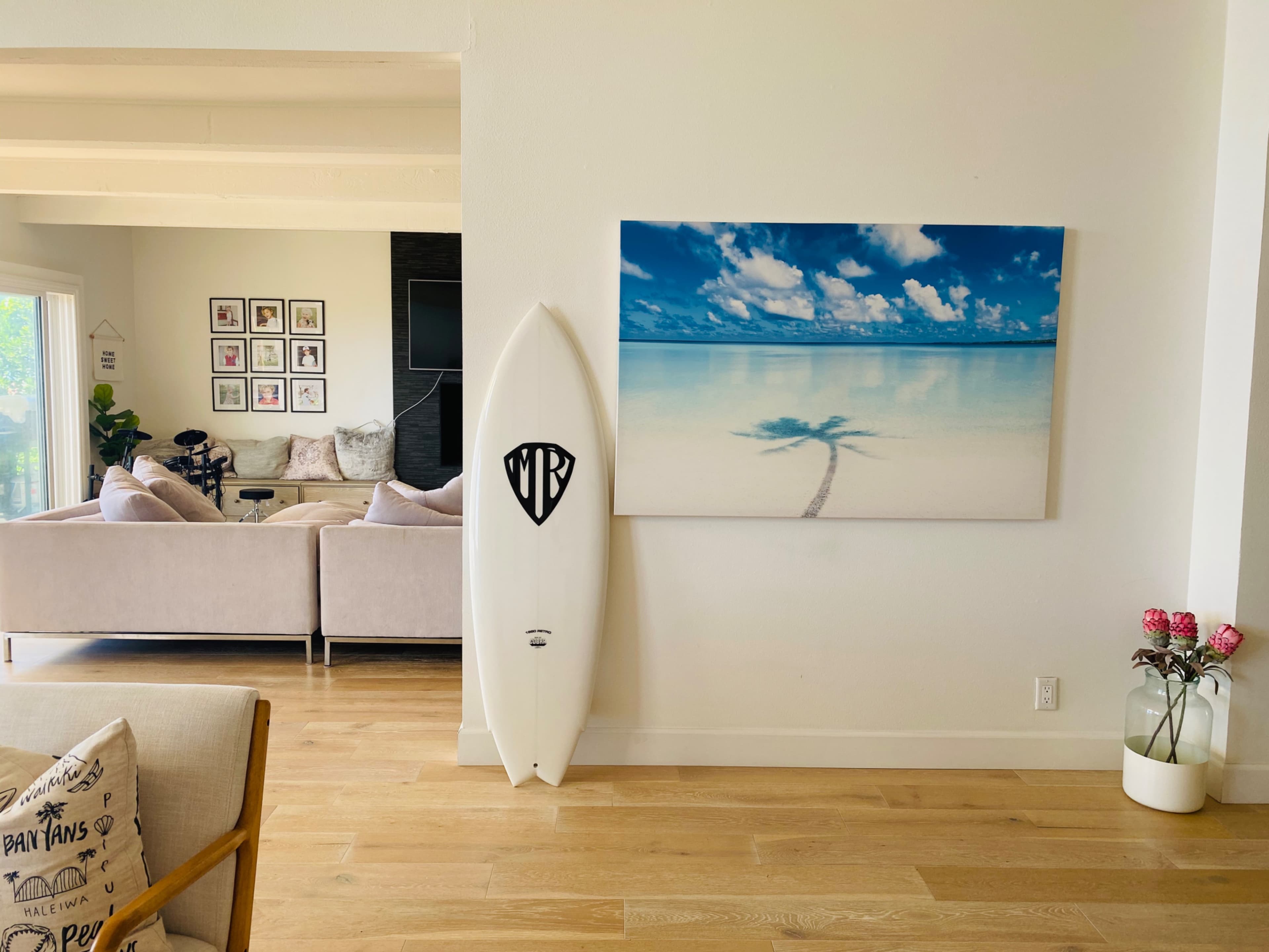 A white surfboard stands against a wall beside a large photograph of a serene beach scene.