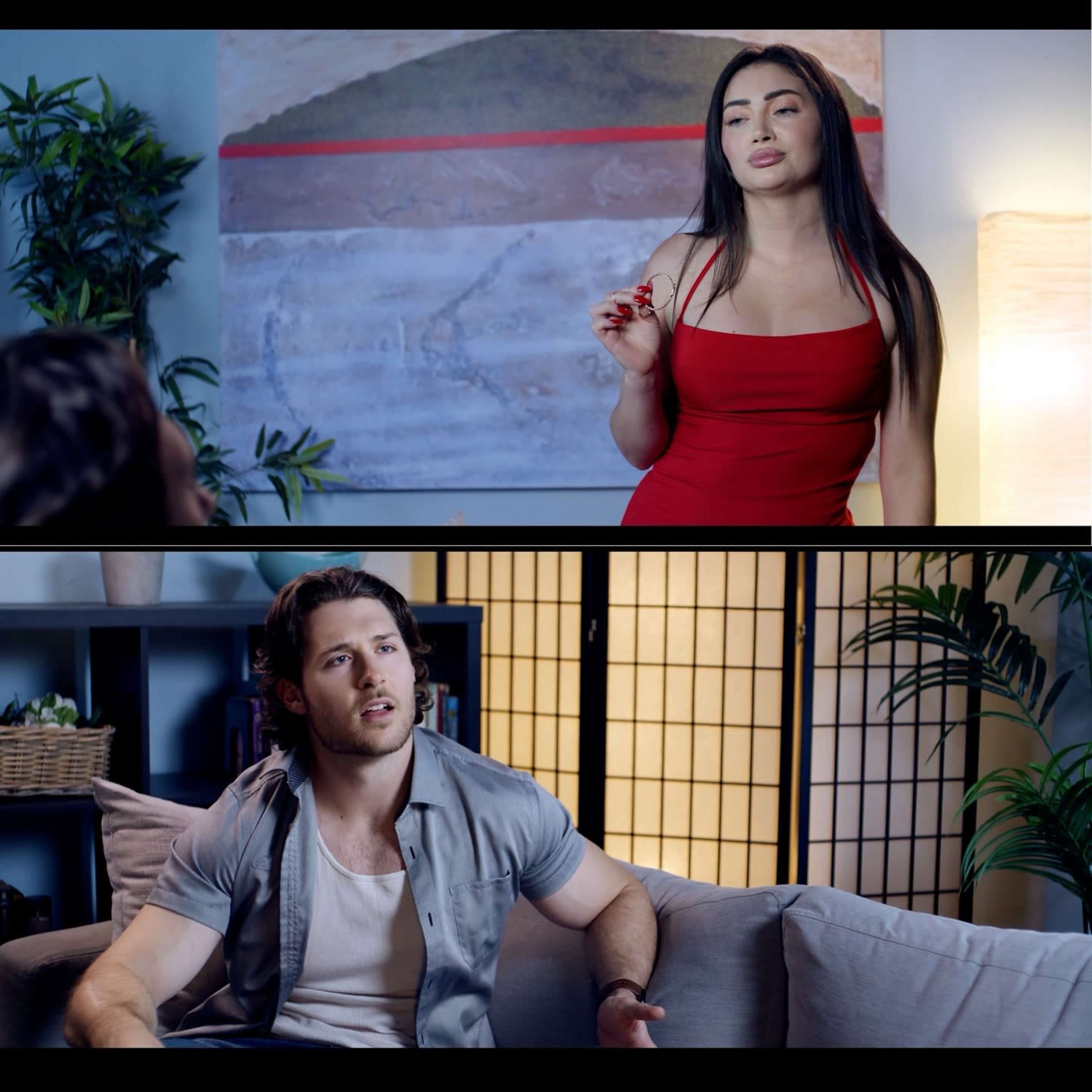 A woman in a red dress stands while holding a drink, and a man in a gray shirt sits on a couch, looking at her in a well-lit room.