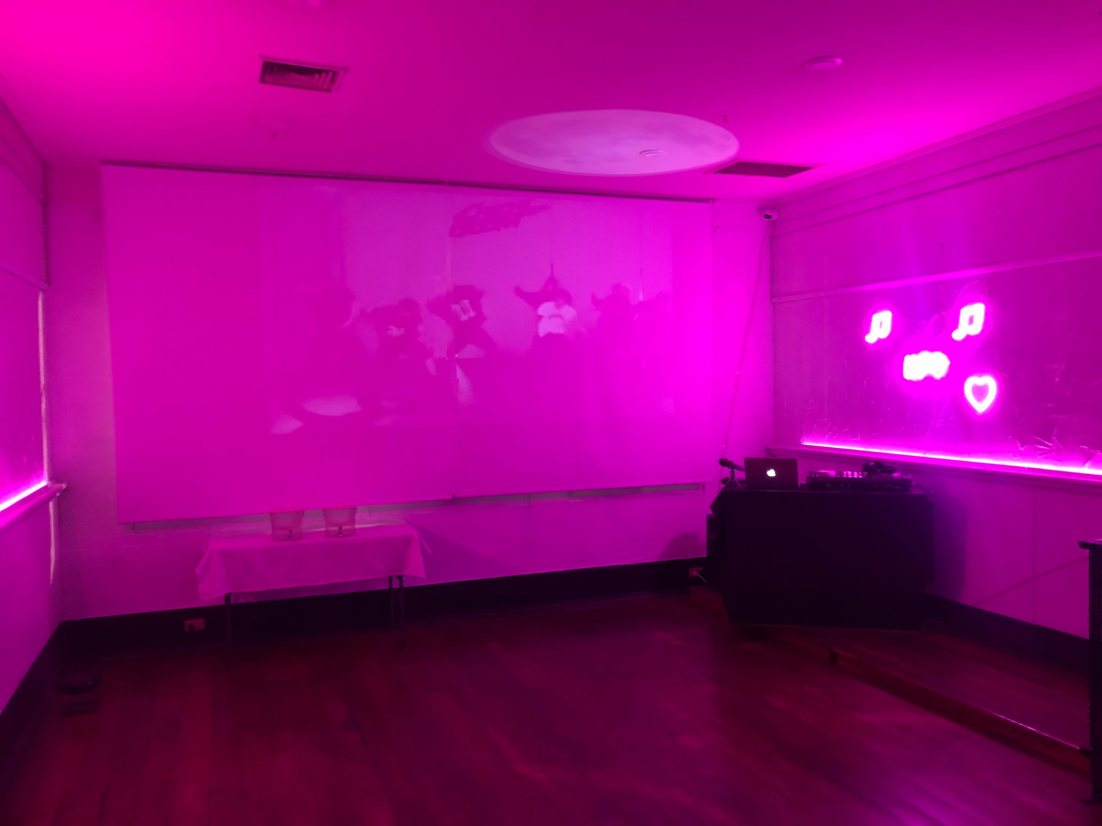 The room features pink ambient lighting, a projector screen at the front, and a music setup on a black table.