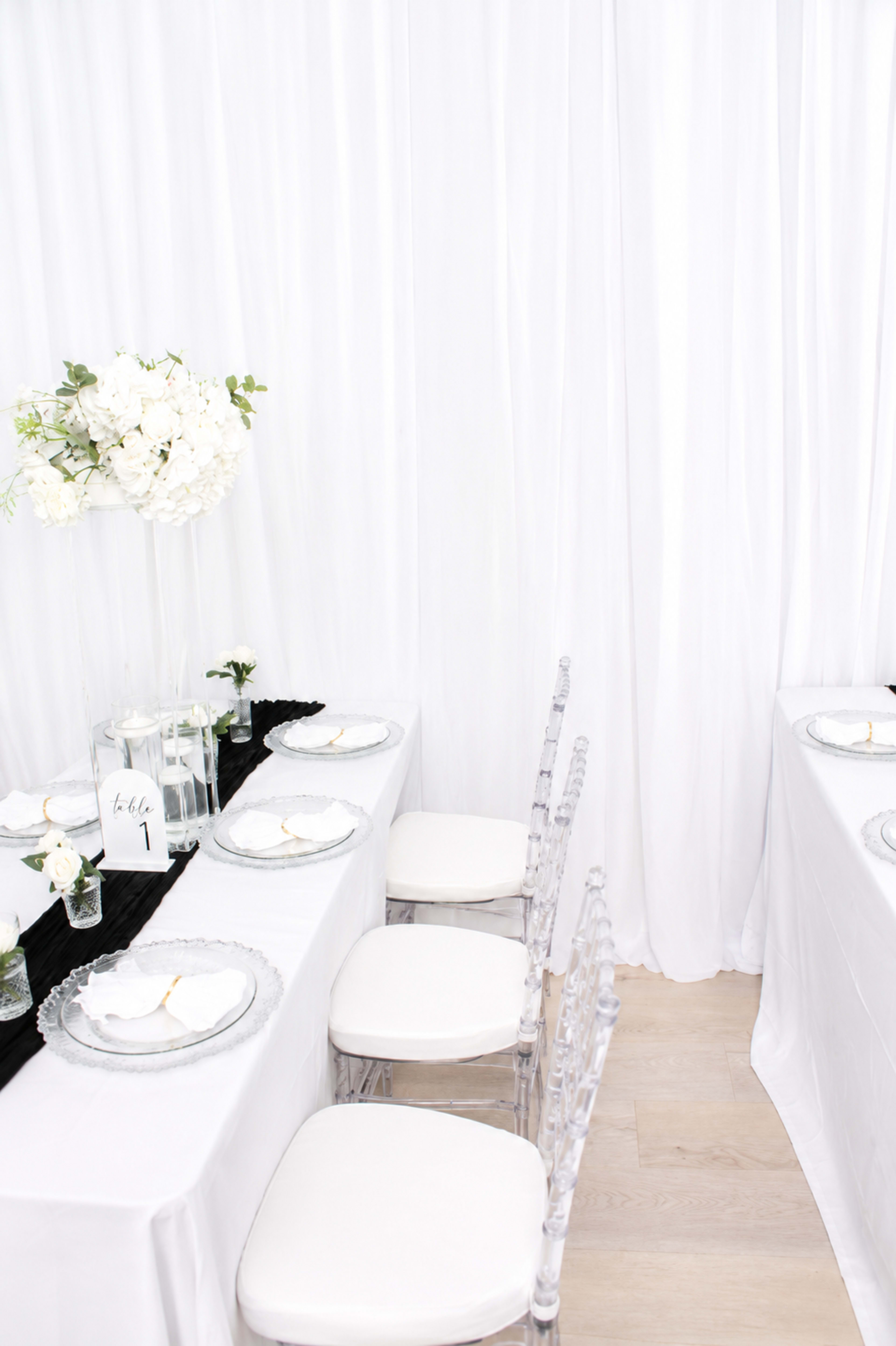 The image shows a dining setup with two white tables adorned with elegant tableware, crystal chairs, and floral centerpieces against a backdrop of white drapery.